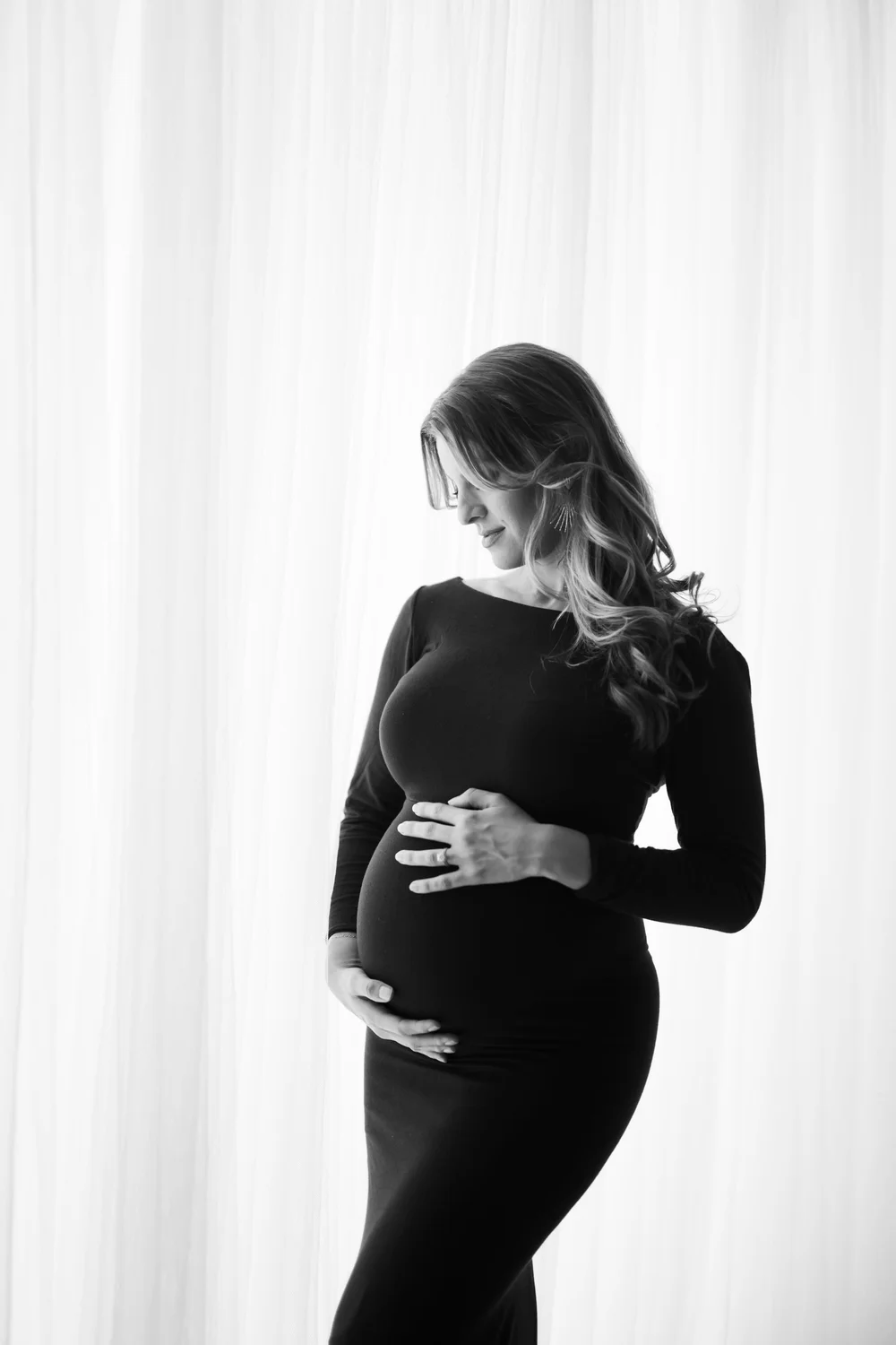 Black-and-white maternity portrait beside softly lit curtains