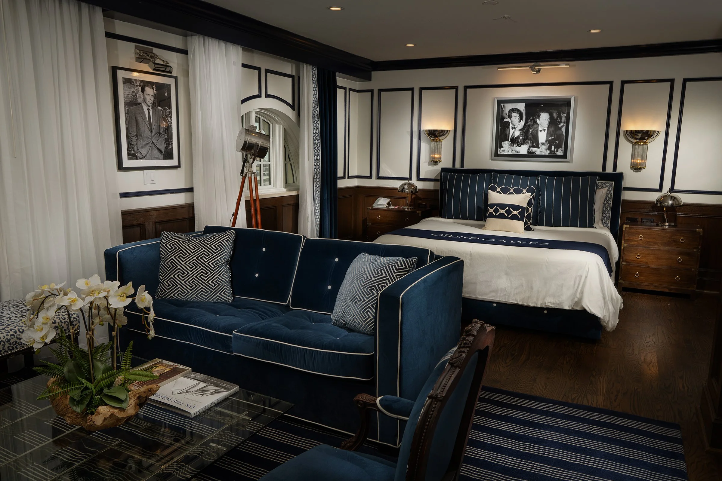 A luxurious hotel room featuring a large bed with navy blue and white bedding, framed black and white photographs on the walls, a navy blue velvet sofa with patterned pillows, a glass-top coffee table with a flower arrangement, and elegant wall light