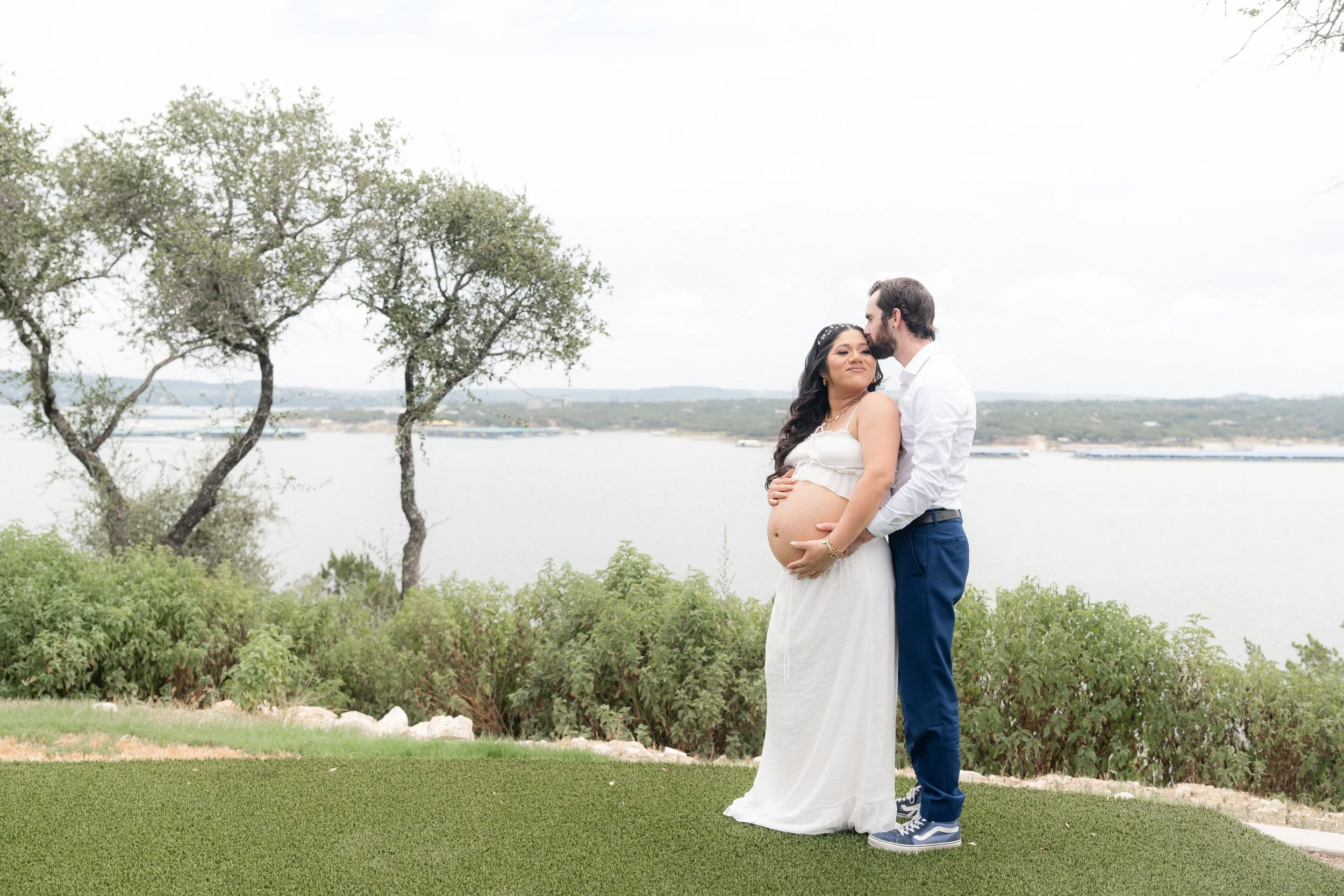 A pregnant woman in a white dress and a man in a white shirt and blue pants standing outdoors near water, with trees and greenery in the background. The man is kissing the woman on her forehead while she gently rests her hand on her belly.