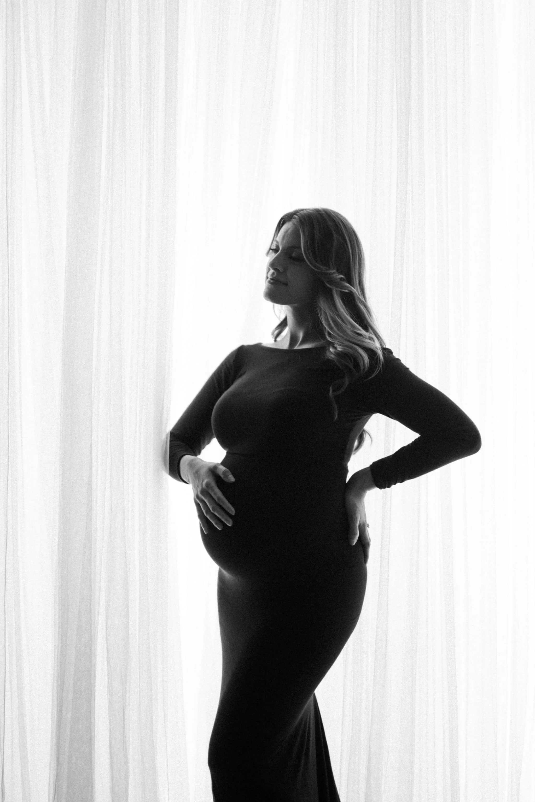 A pregnant woman in a long black dress standing in front of a sheer white curtain. She has styled hair and is posing with her left hand on her hip and her right hand resting gently on her belly.