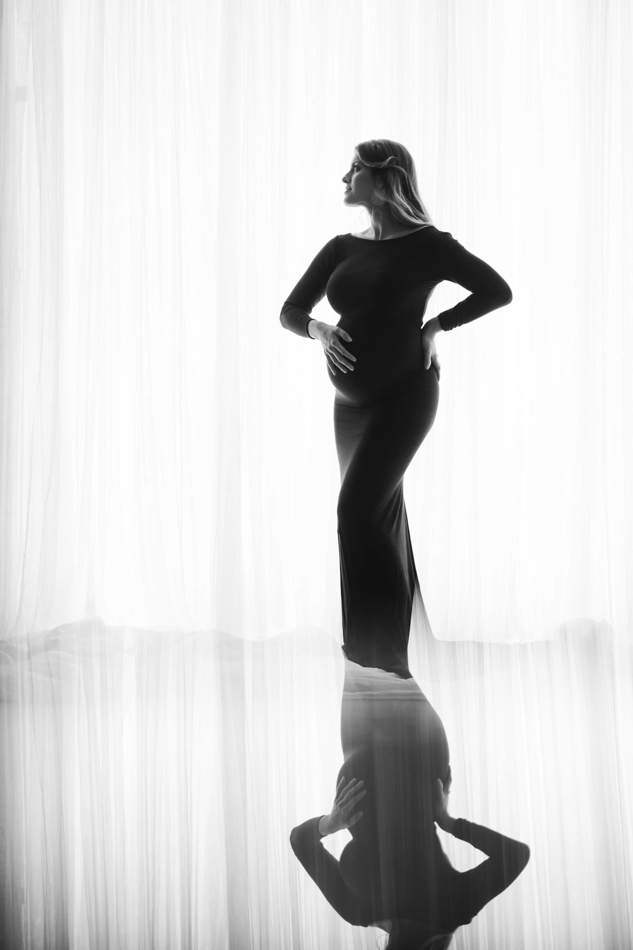 Black and white photo of a woman standing with her hands on her hips in front of a light-colored curtain, with her reflection visible underneath.