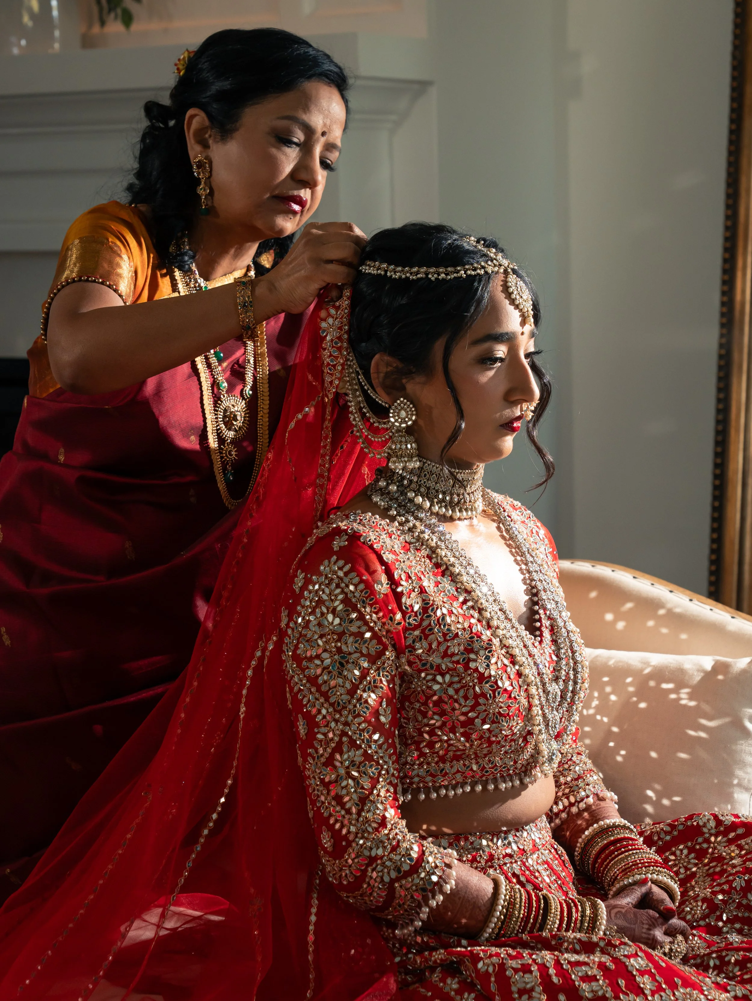 Indian bride dressed in red and gold bridal attire with intricate jewelry, sitting as a woman adjusts her headpiece at the Videre Estate.
