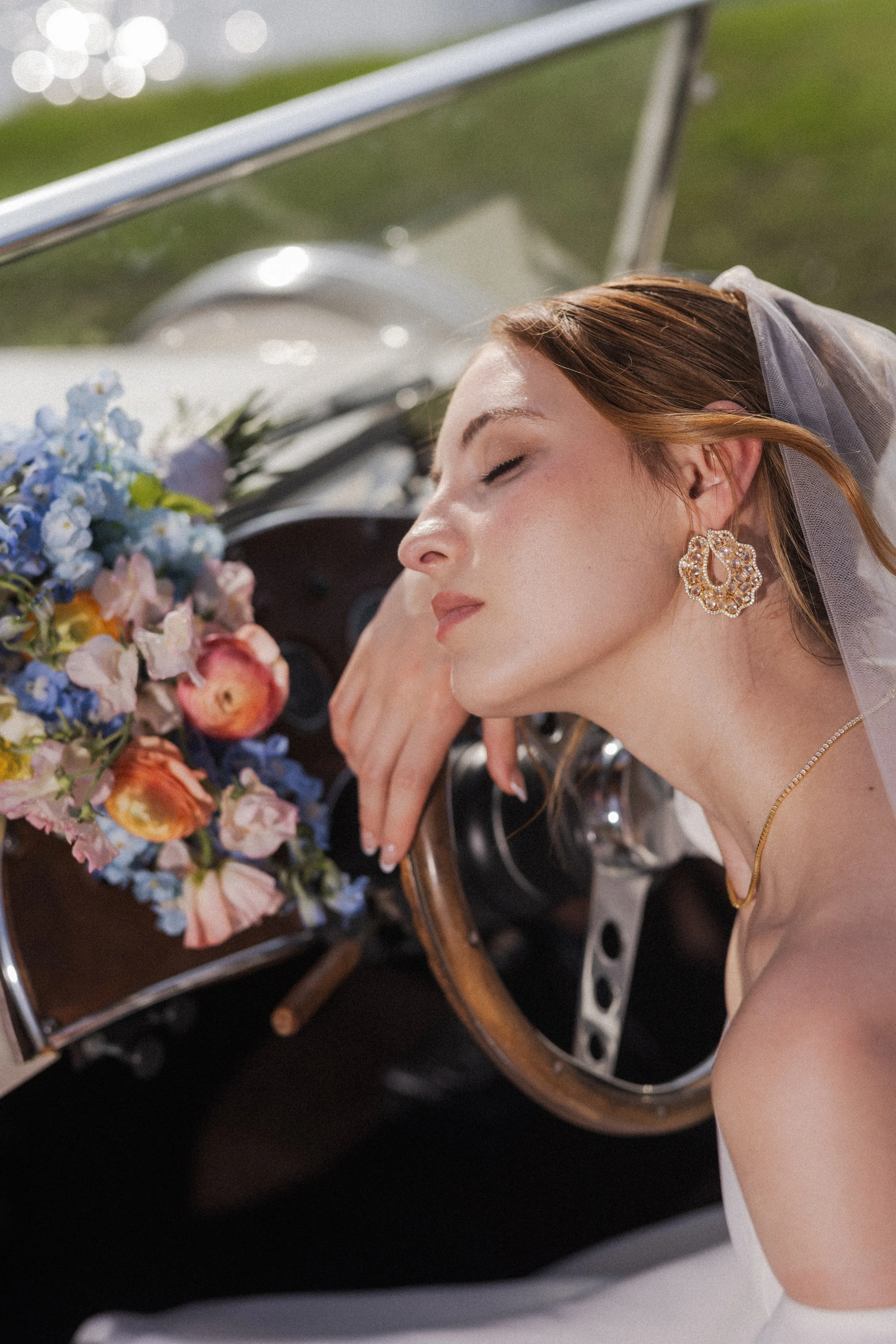 A bride with red hair, closed eyes, and gold earrings leaning on a vintage car with a bouquet of colorful flowers on the dashboard at Ma Maison wedding venue.