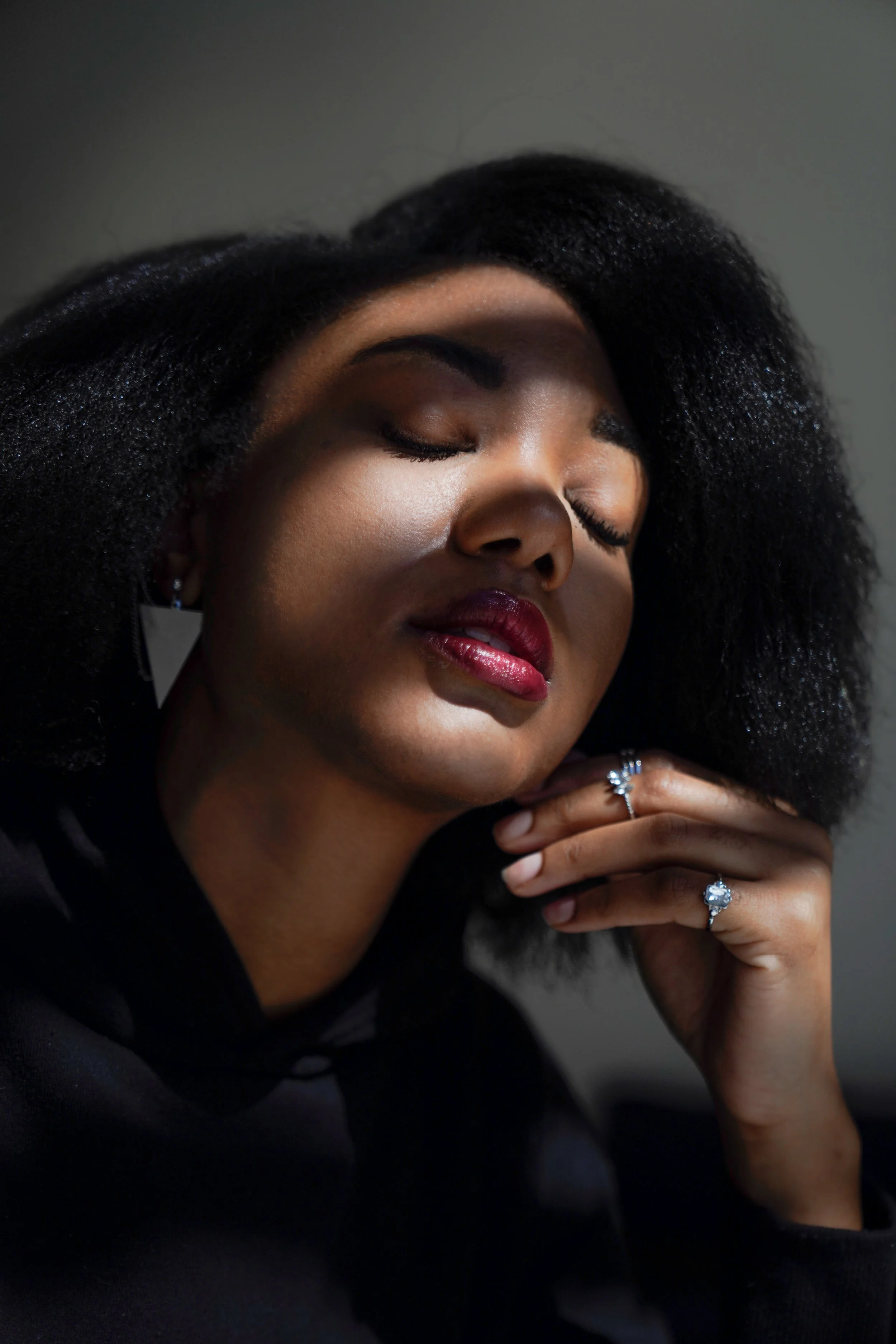 Close-up of a woman with closed eyes, wearing dark makeup and a ring on her finger, resting her hand near her chin.
