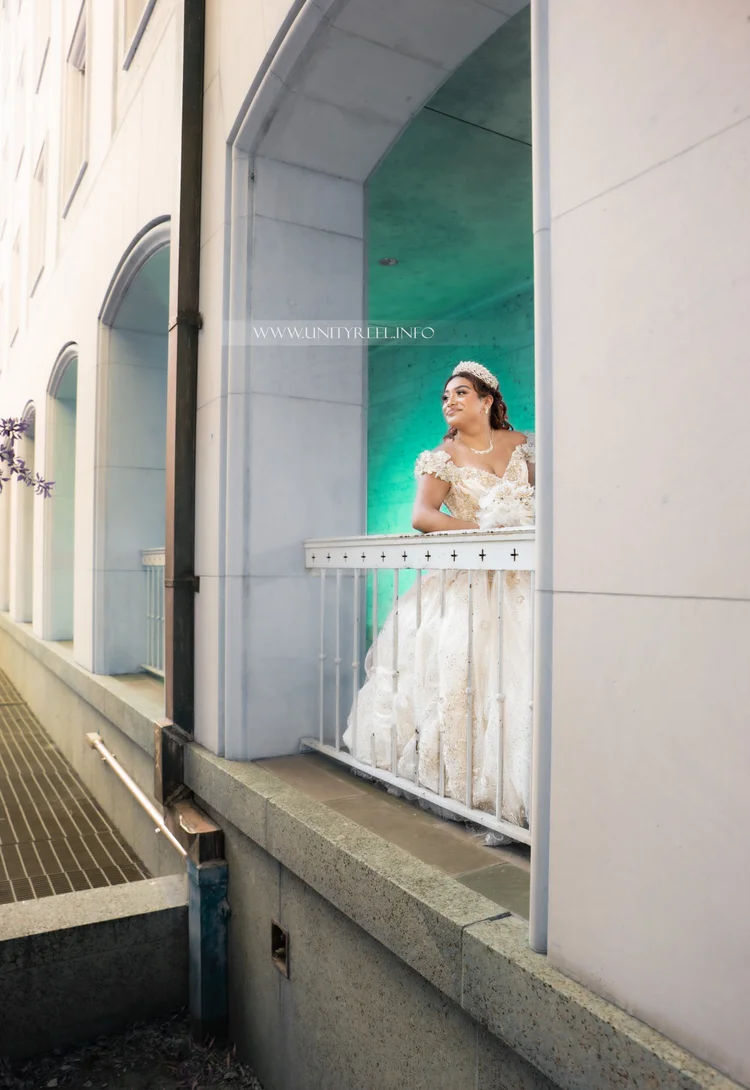 Portrait in a white gown and tiara on a balcony, smiling toward the view.