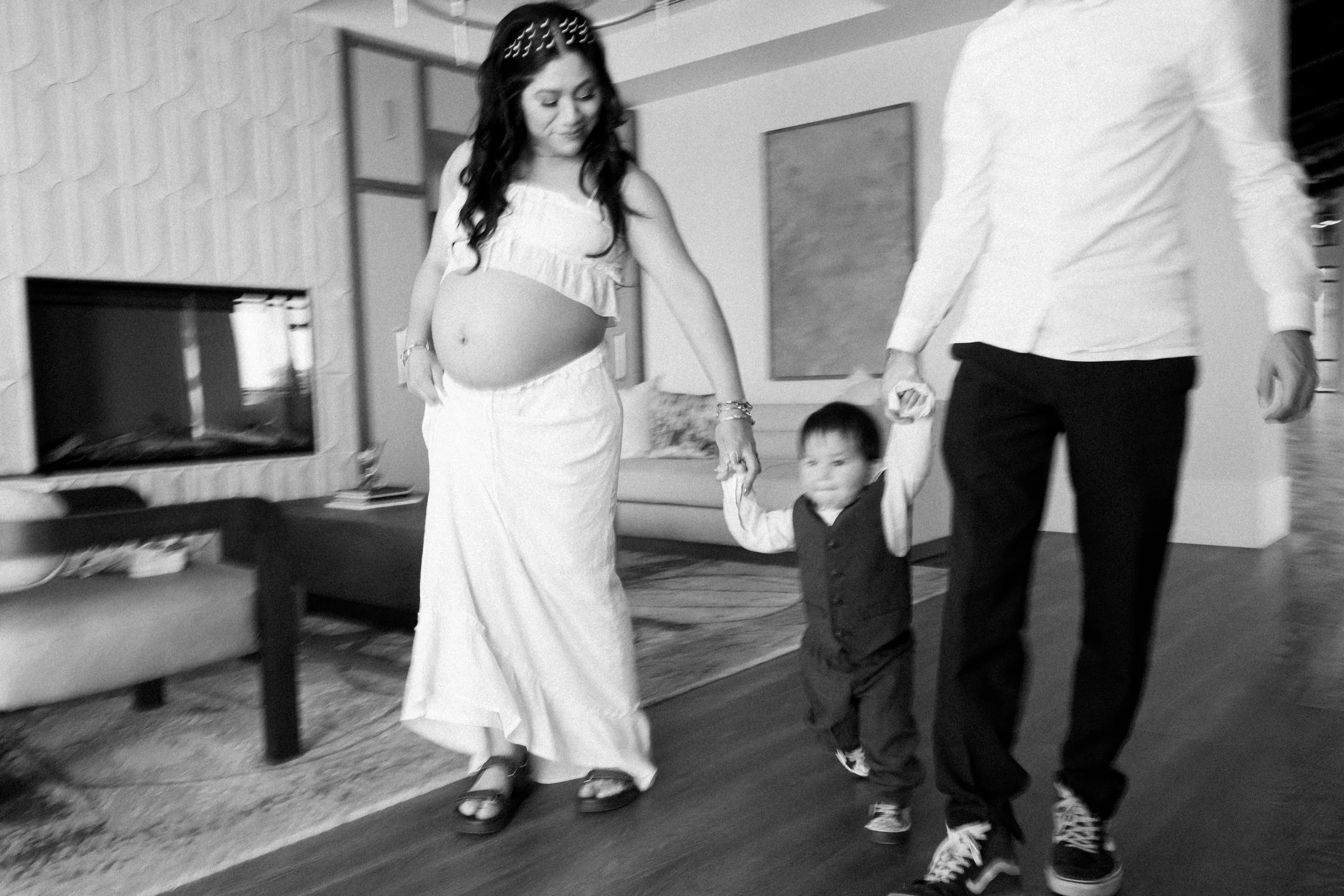A pregnant woman in a white dress holding hands with a young child, walking indoors with a man in a white shirt and black pants.