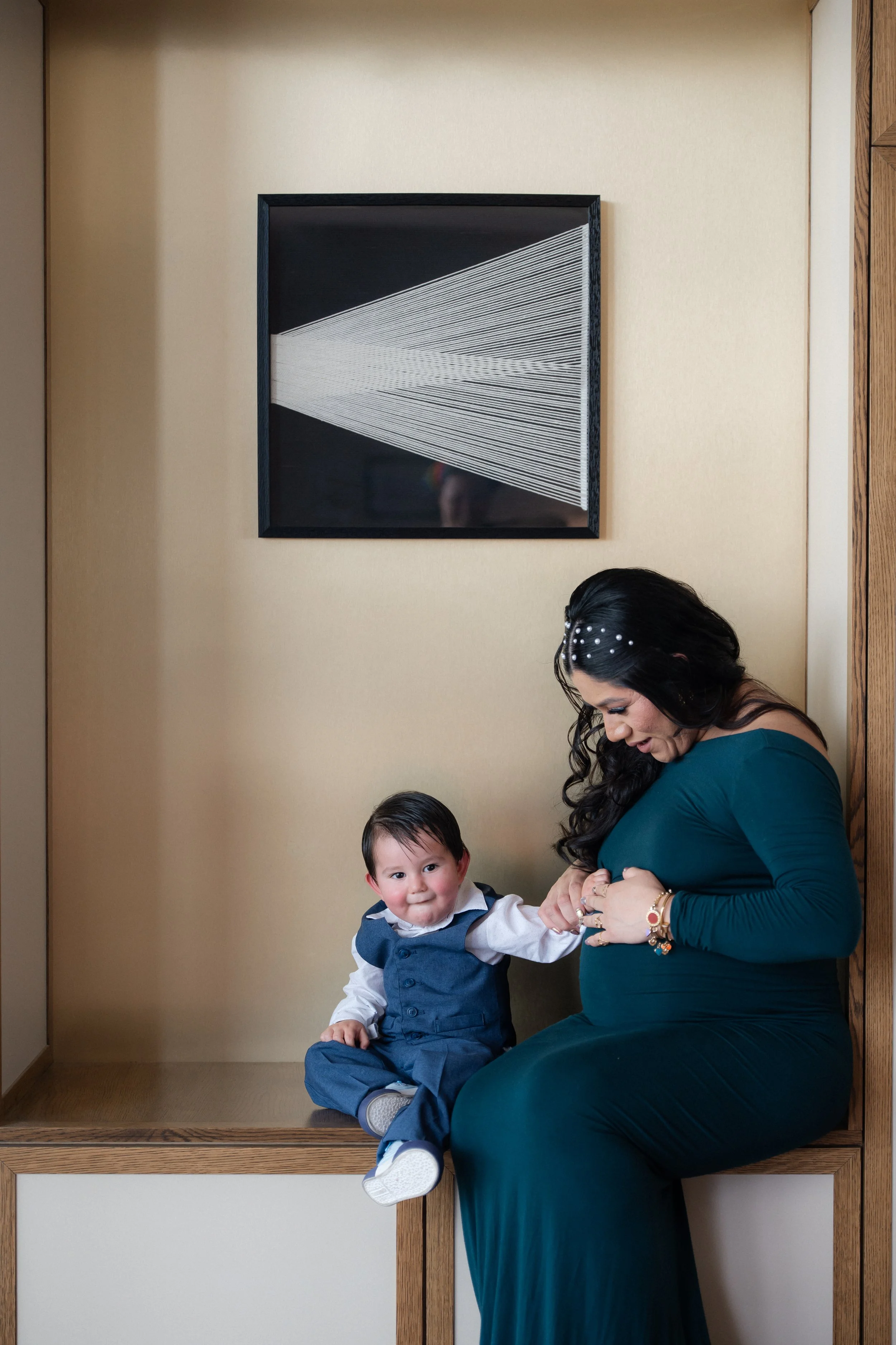 A woman with long dark hair and a dark teal dress is sitting on a wooden ledge, holding a young boy in a blue vest and gray pants. They are in front of a beige wall with a geometric black and white artwork hanging above. The woman is smiling and the 