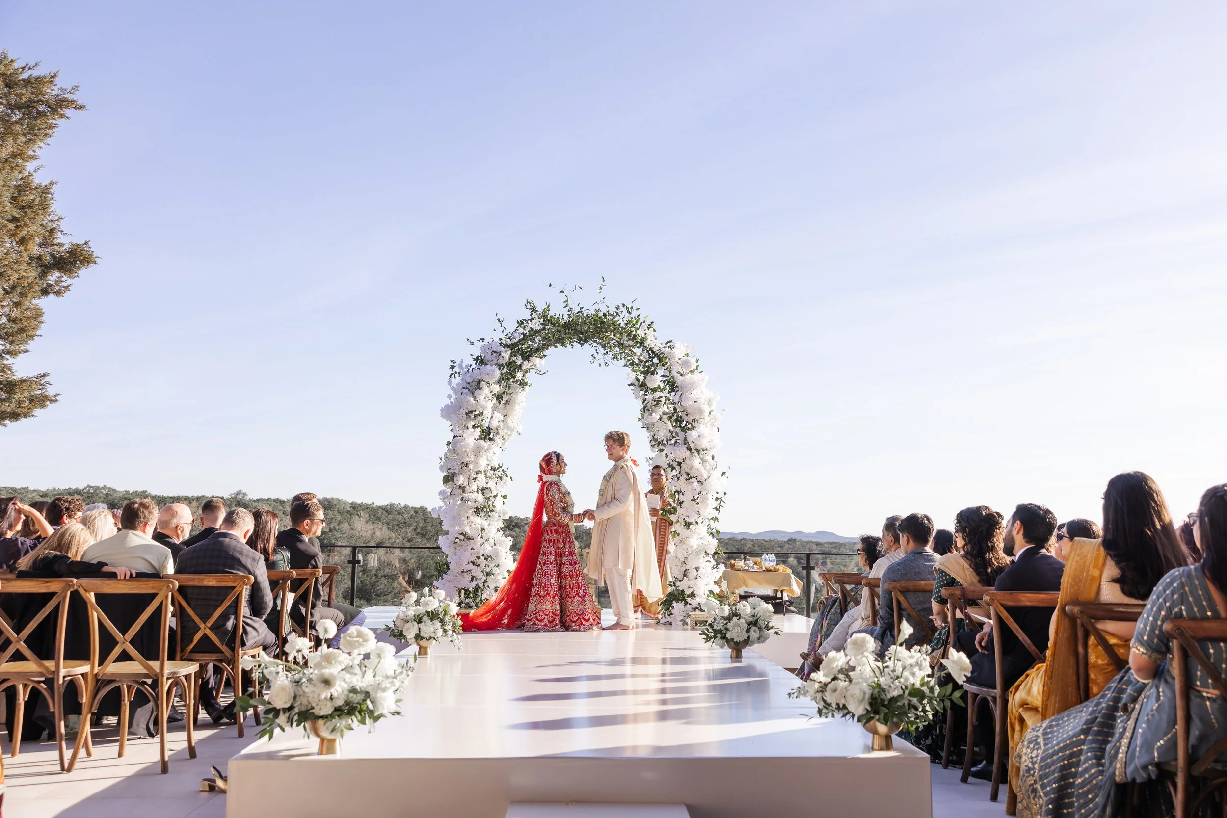 An outdoor wedding ceremony with a bride and groom exchanging vows under a floral arch, surrounded by seated guests on a bright, clear day on the terrace at the Videre Estate.