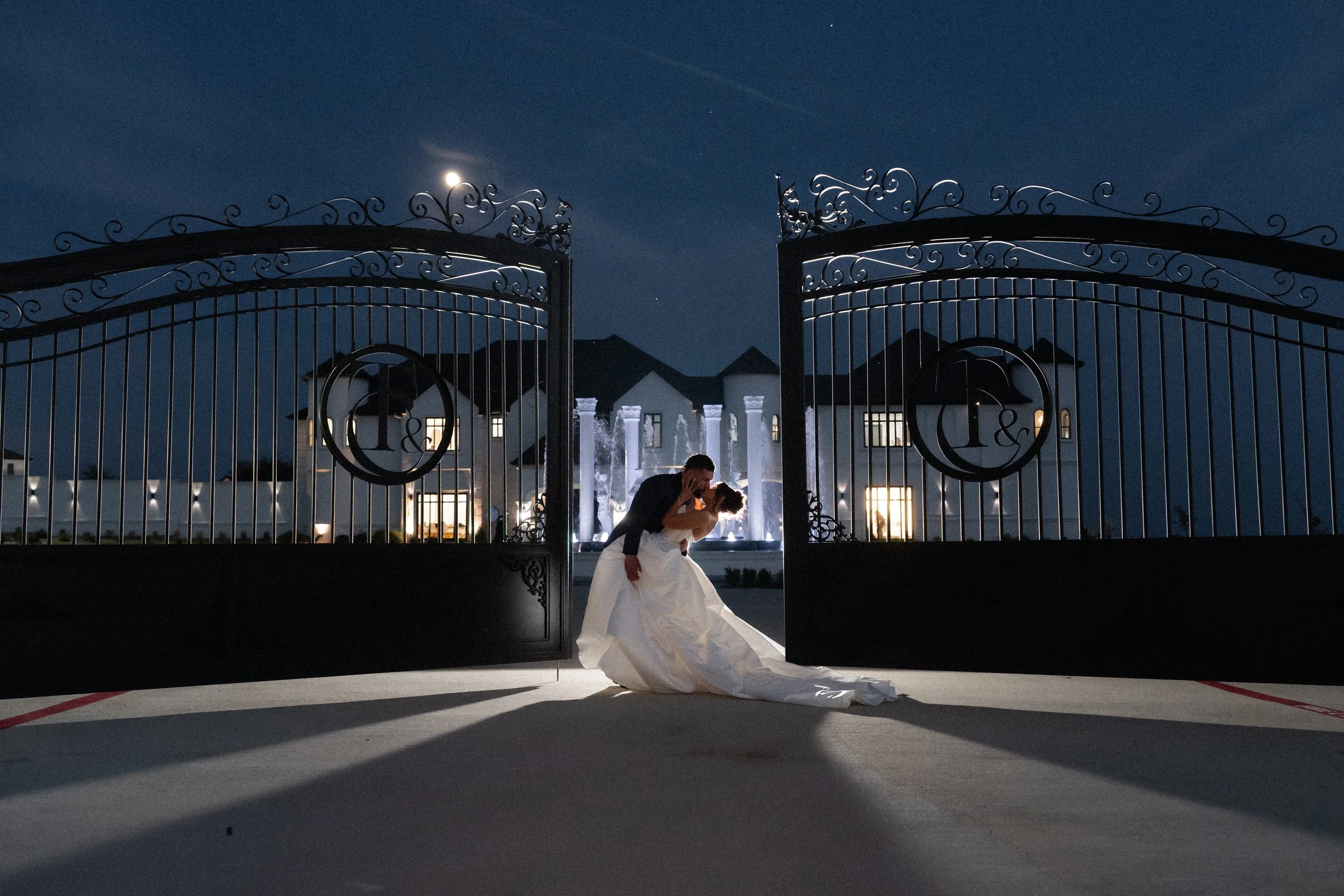 Bride and groom kissing beneath an ornate estate gate at night.