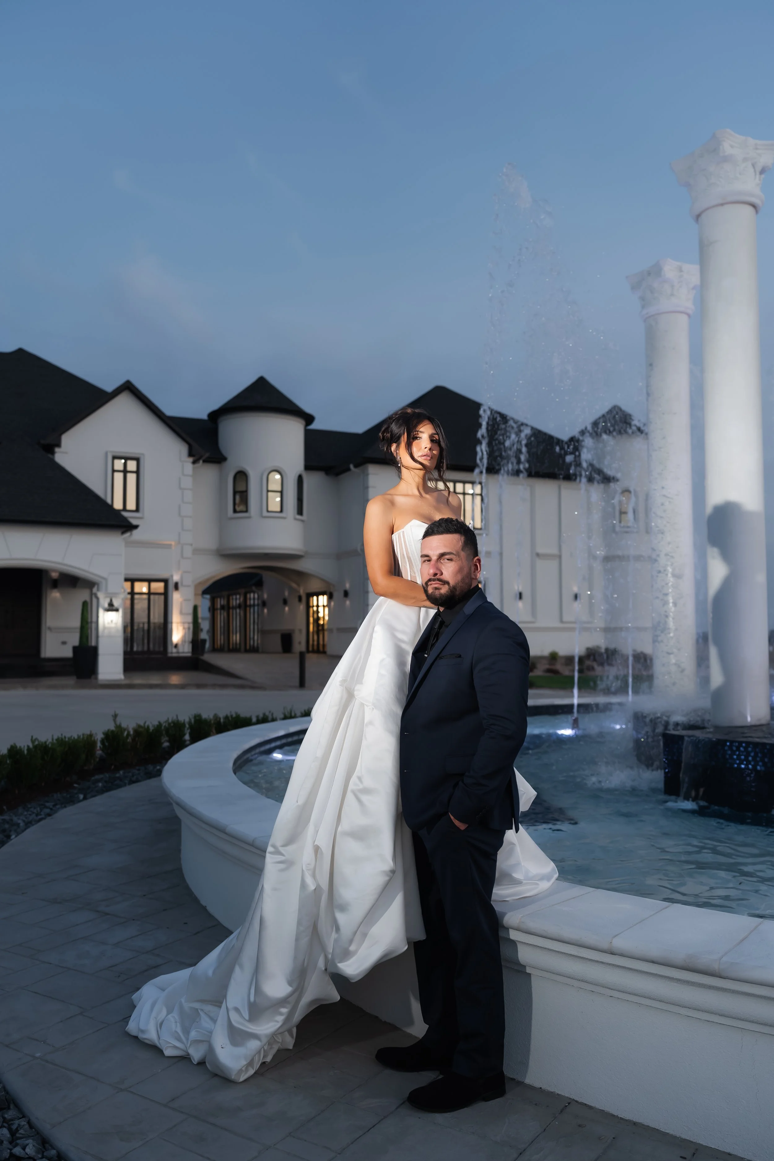 Newlyweds posing beside a fountain outside a white estate at dusk.