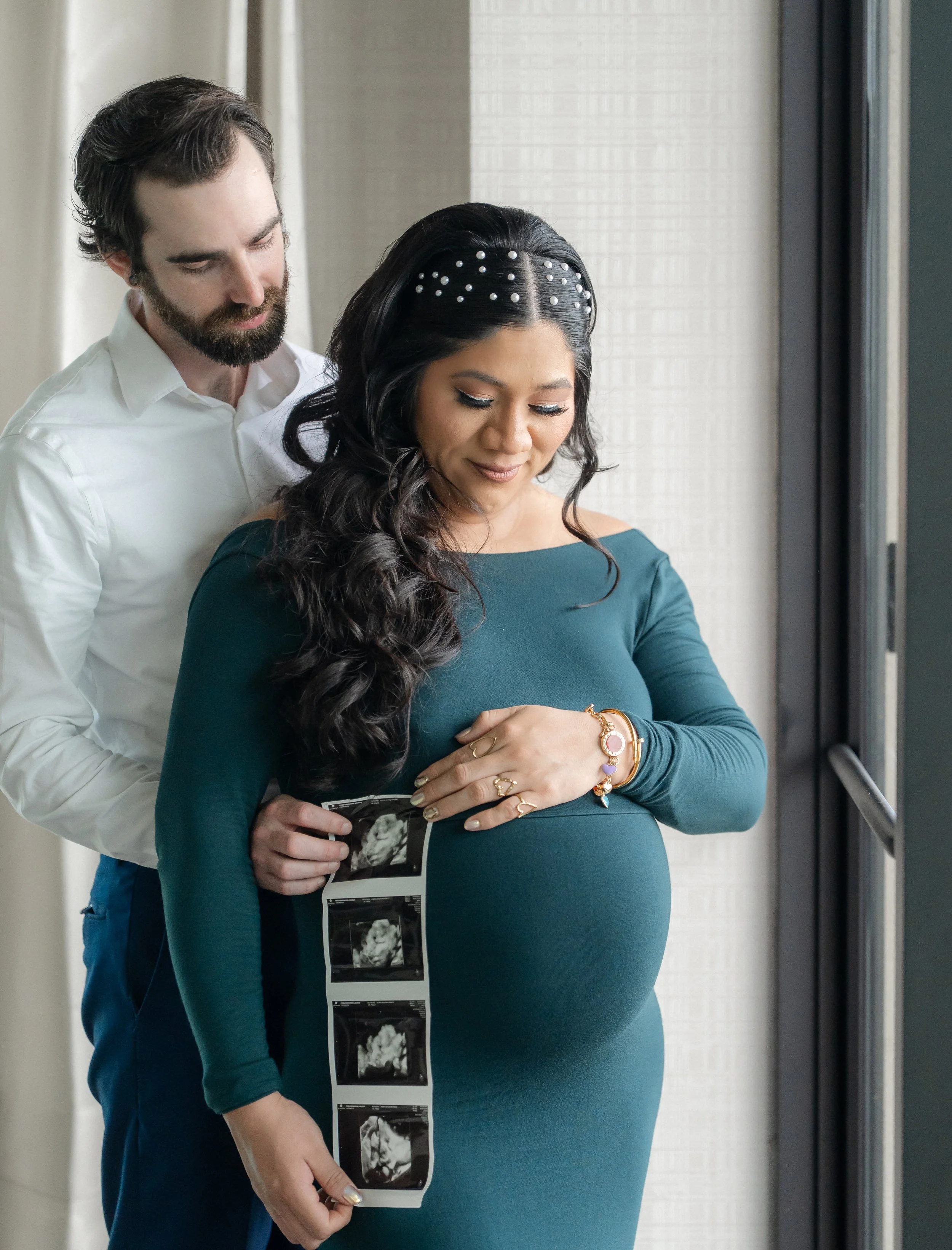 A pregnant woman holding ultrasound images, standing in an elevator, with a man behind her looking at the ultrasound pictures.