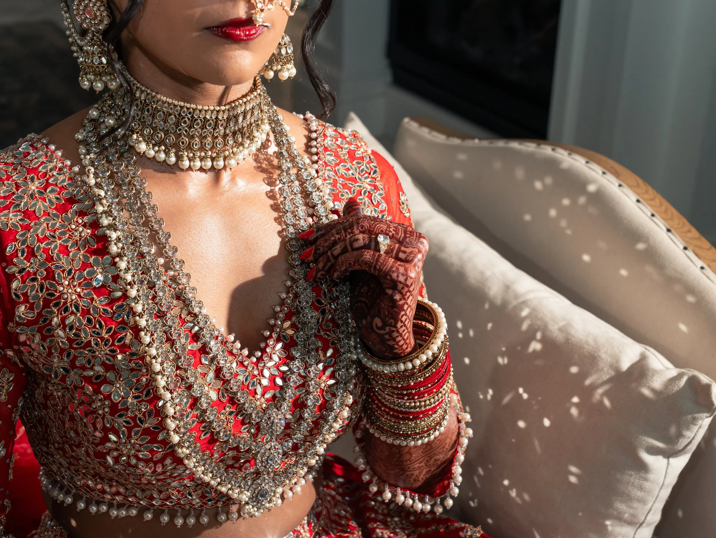 Close-up of a bride in traditional Indian attire, adorned with elaborate jewelry including necklaces, earrings, and bangles, wearing a red embroidered outfit, with henna decorated hands, sitting in the bridal suite at the Videre Estate.