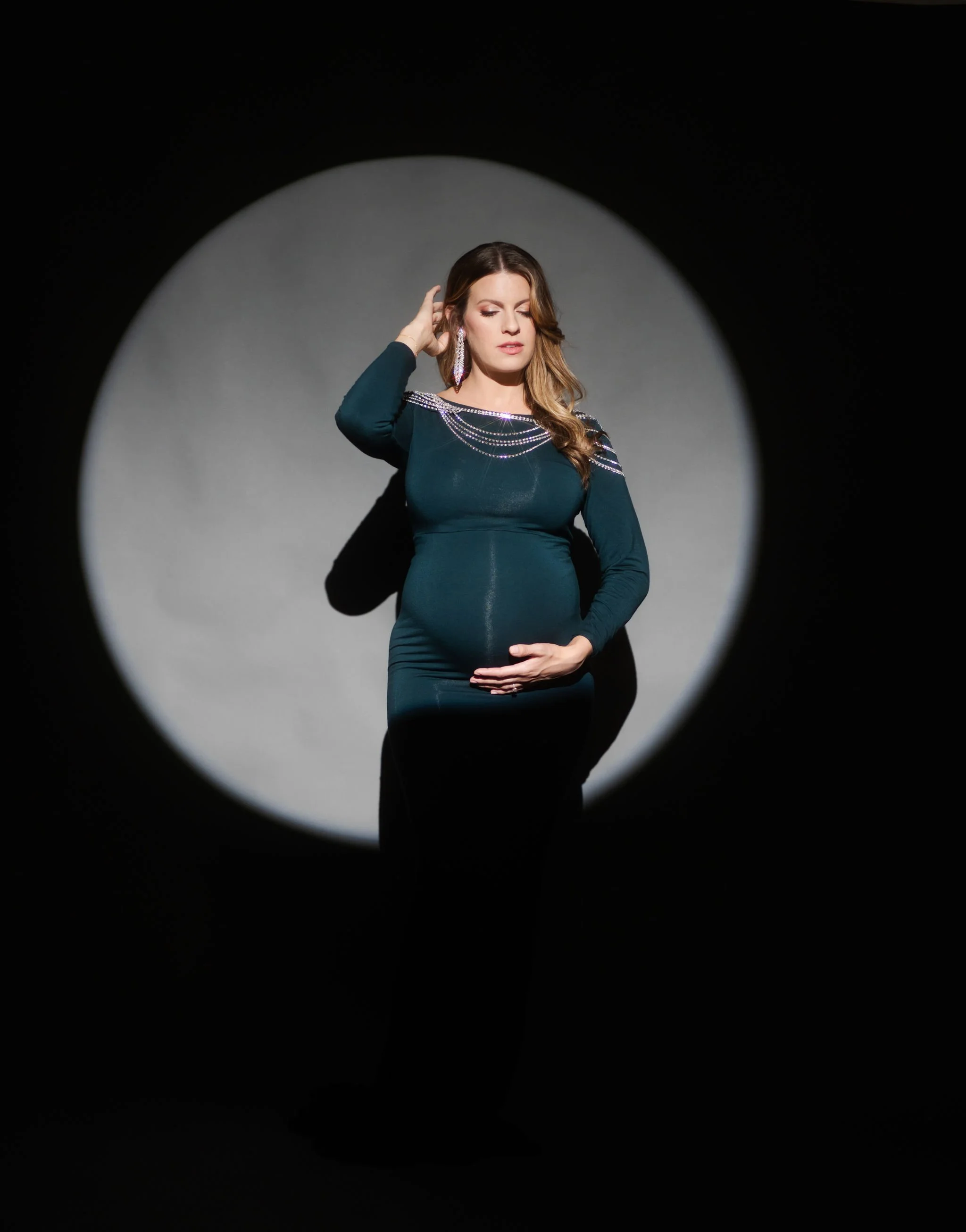 A pregnant woman with long wavy hair and earrings, wearing a dark teal dress with rhinestone embellishments on the shoulders, posing with a closed eyes, sincere expression, and hand on her baby bump, against a gray backdrop, framed within a circular 