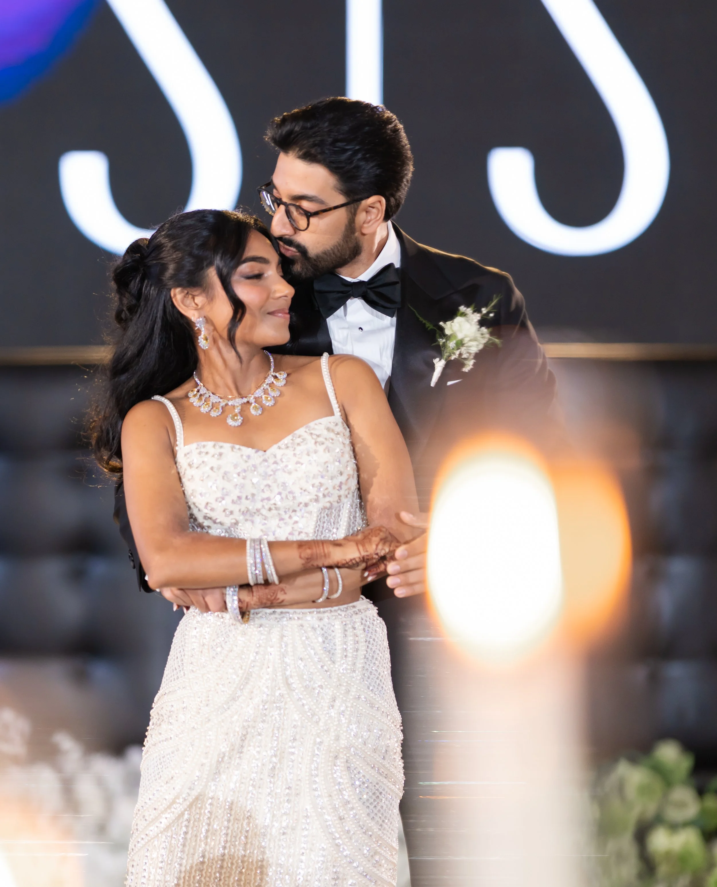 A newlywed Indian couple sharing an intimate dance at their wedding reception. The bride wears a white lace gown and jewelry, and the groom is dressed in a tuxedo with a bow tie and boutonniere. They are embracing with their eyes closed.