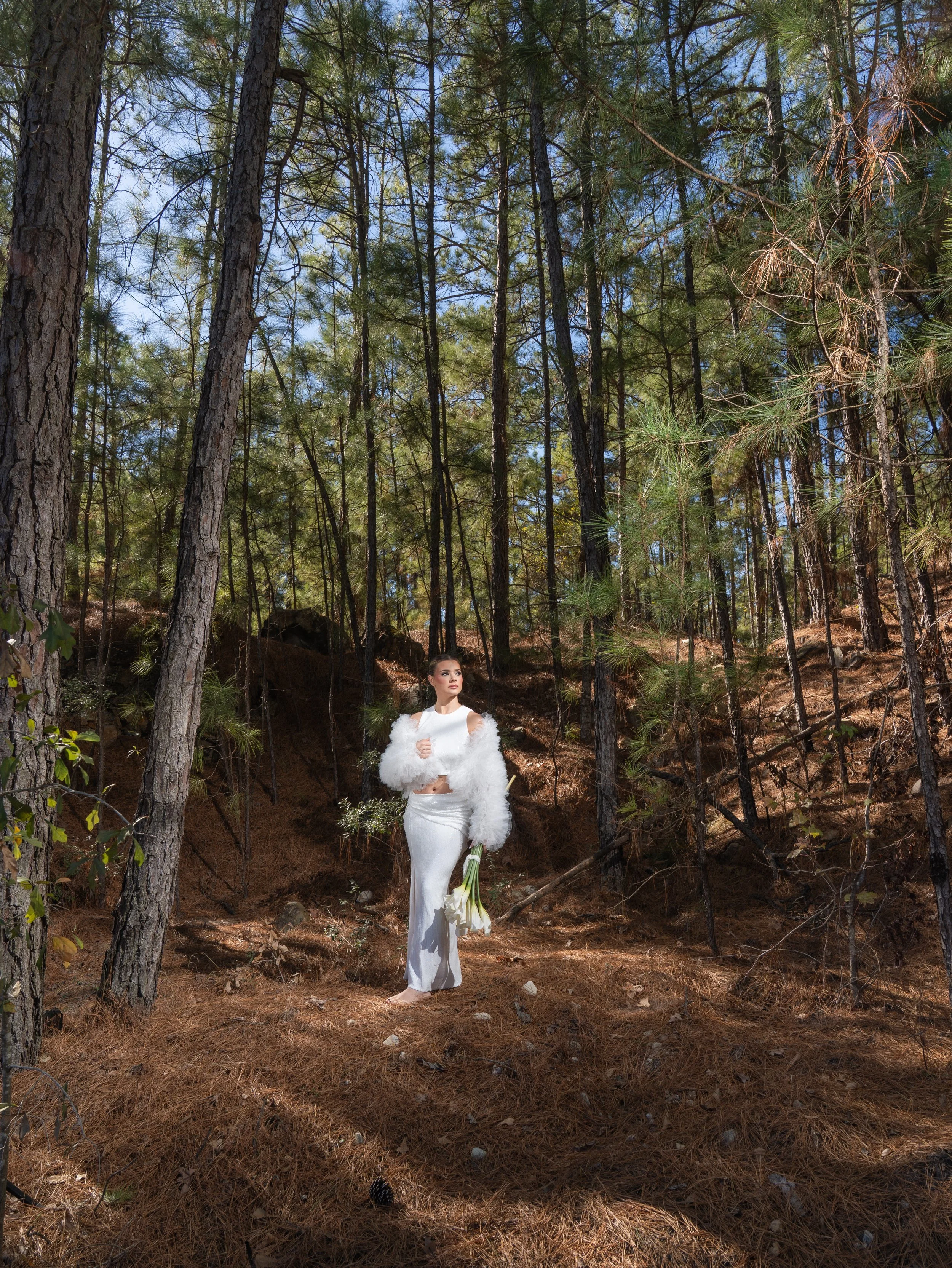 A woman dressed in a white gown and fur coat stands in a forest surrounded by tall pine trees, holding a bouquet of white calla lilies from an elegant editorial wedding styled shoot in Houston.