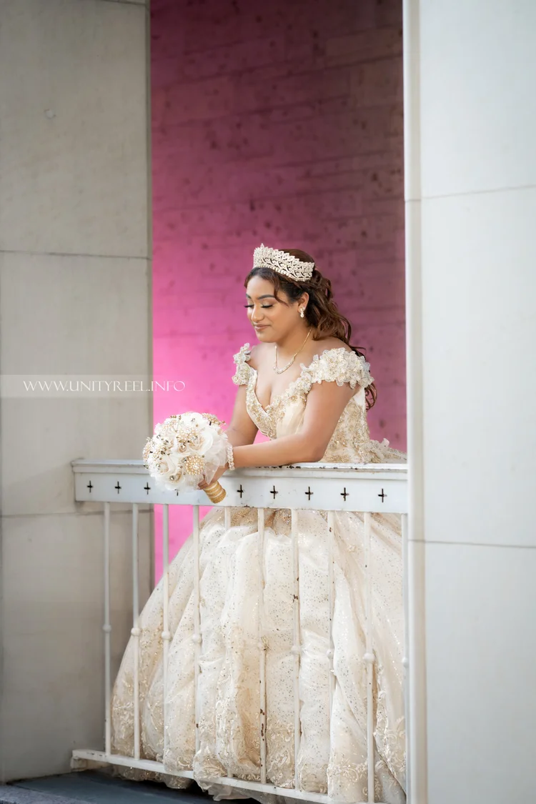Portrait in an ornate cream gown and tiara, holding a bouquet beside a white balcony railing.