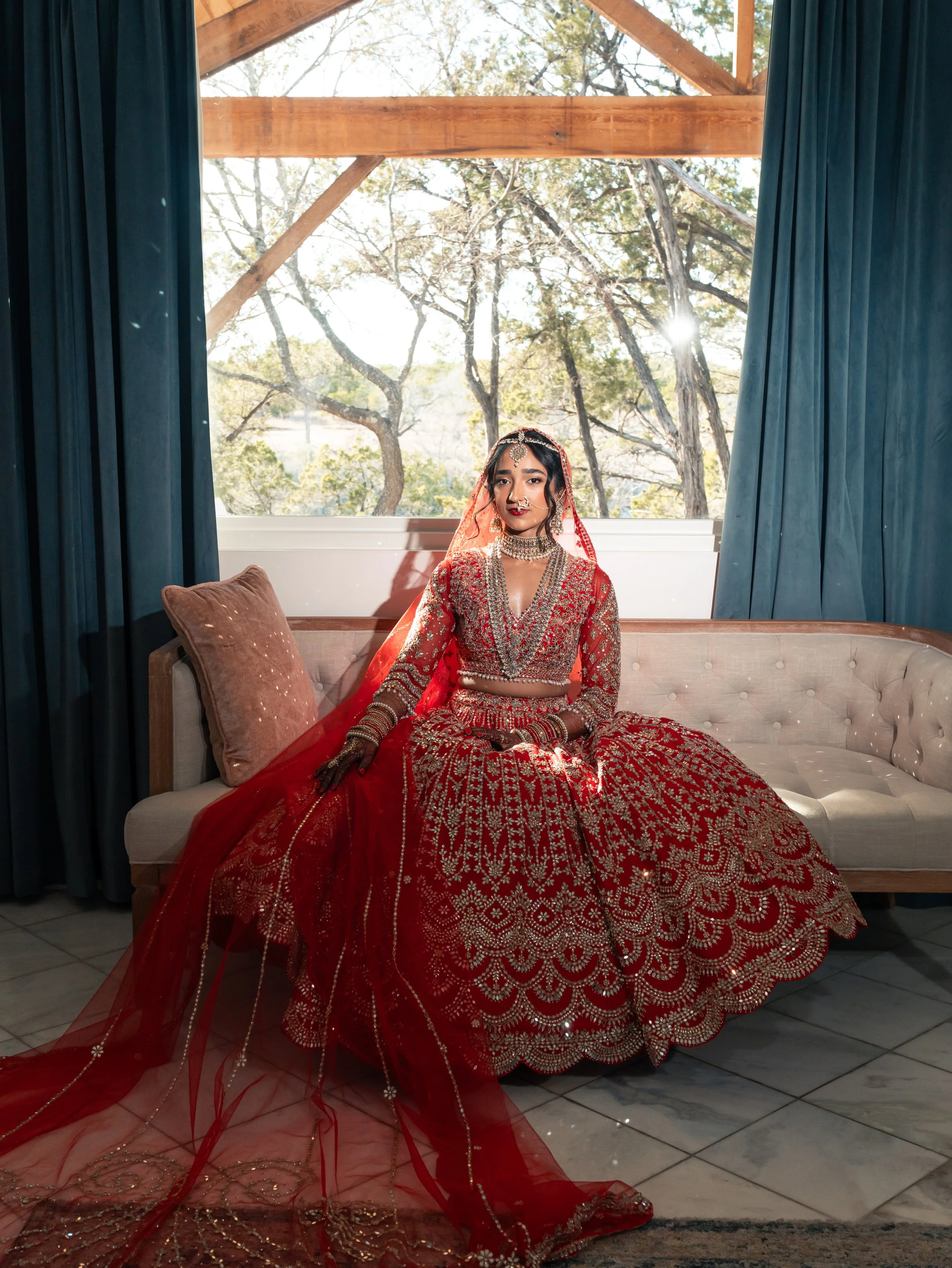 A woman in traditional red and gold wedding attire sitting on a beige couch with blue curtains and a large window showing trees and sunlight in the background in the bridal suite at the Videre Estate.