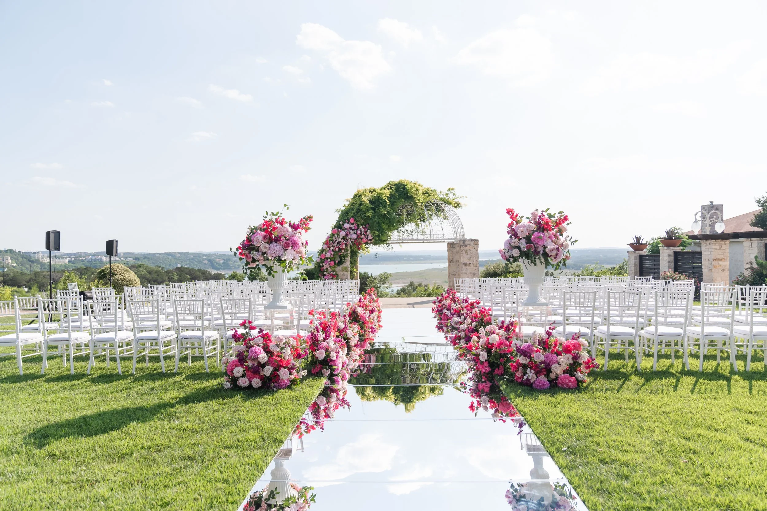 Outdoor wedding ceremony setup with white chairs on either side of a reflective walkway, decorated with pink and purple floral arrangements, leading to a floral arch with greenery, overlooking a scenic landscape under a clear sky.