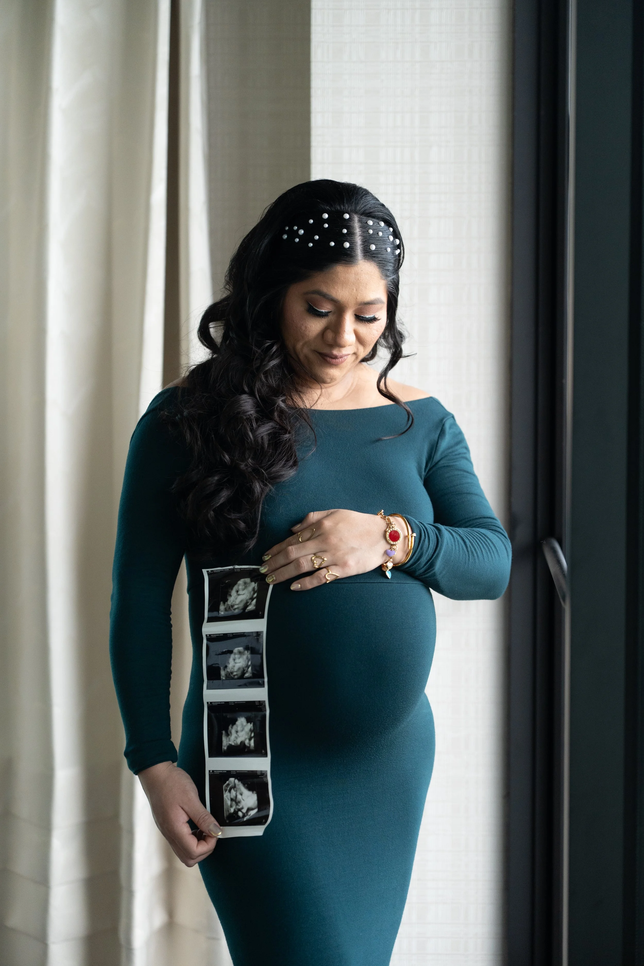A woman with long, dark, curly hair and a headband decorated with pearls stands indoors near a window with curtains. She is wearing a teal, long-sleeved dress and several bracelets, and is looking down at a series of ultrasound images taped to her dr
