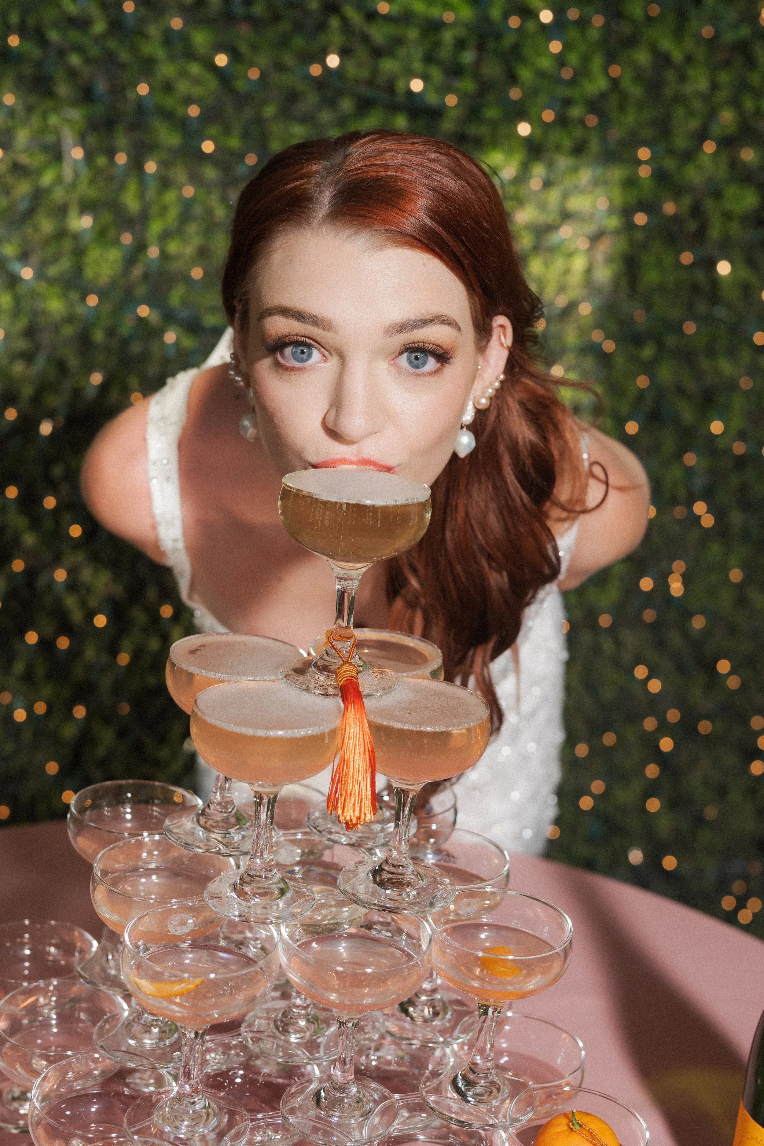 A bride with red hair and pearl earrings leaning over a tower of champagne glasses, possibly at a celebration in the outside reception space at Ma Maison wedding venue.