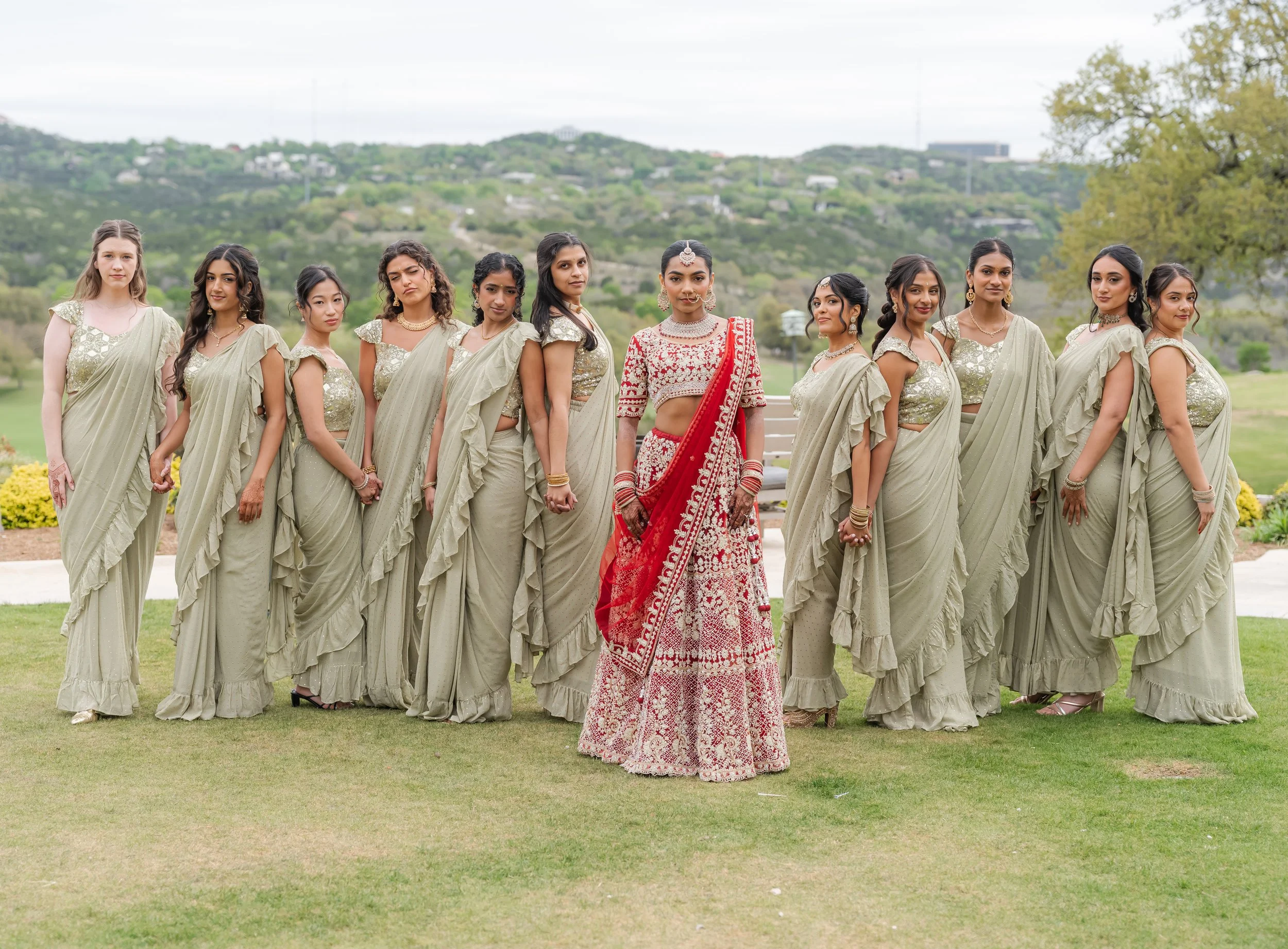 A group of twelve women standing outdoors on a lawn, dressed in traditional Indian attire, with mountains and trees in the background on the greens at the Omni Barton Creek.