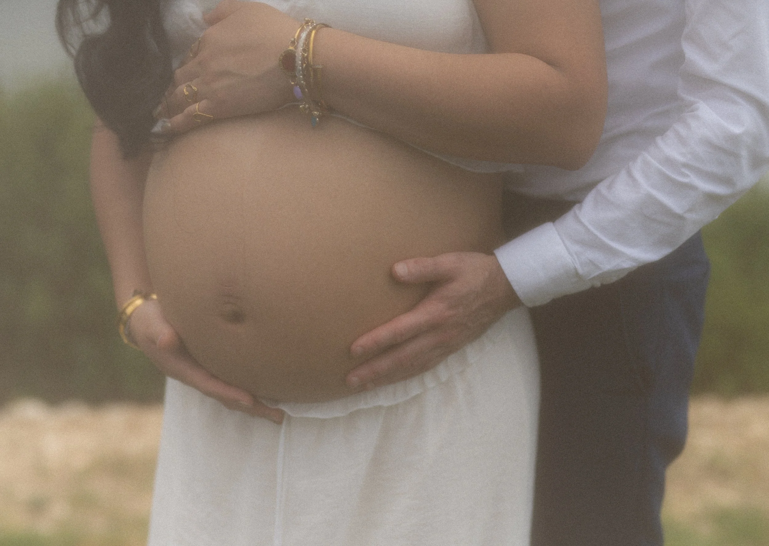 A person holding a pregnant belly outdoors, wearing a white shirt with rolled-up sleeves, with their hands gently cradling the belly. The person is wearing multiple bracelets on their wrist.