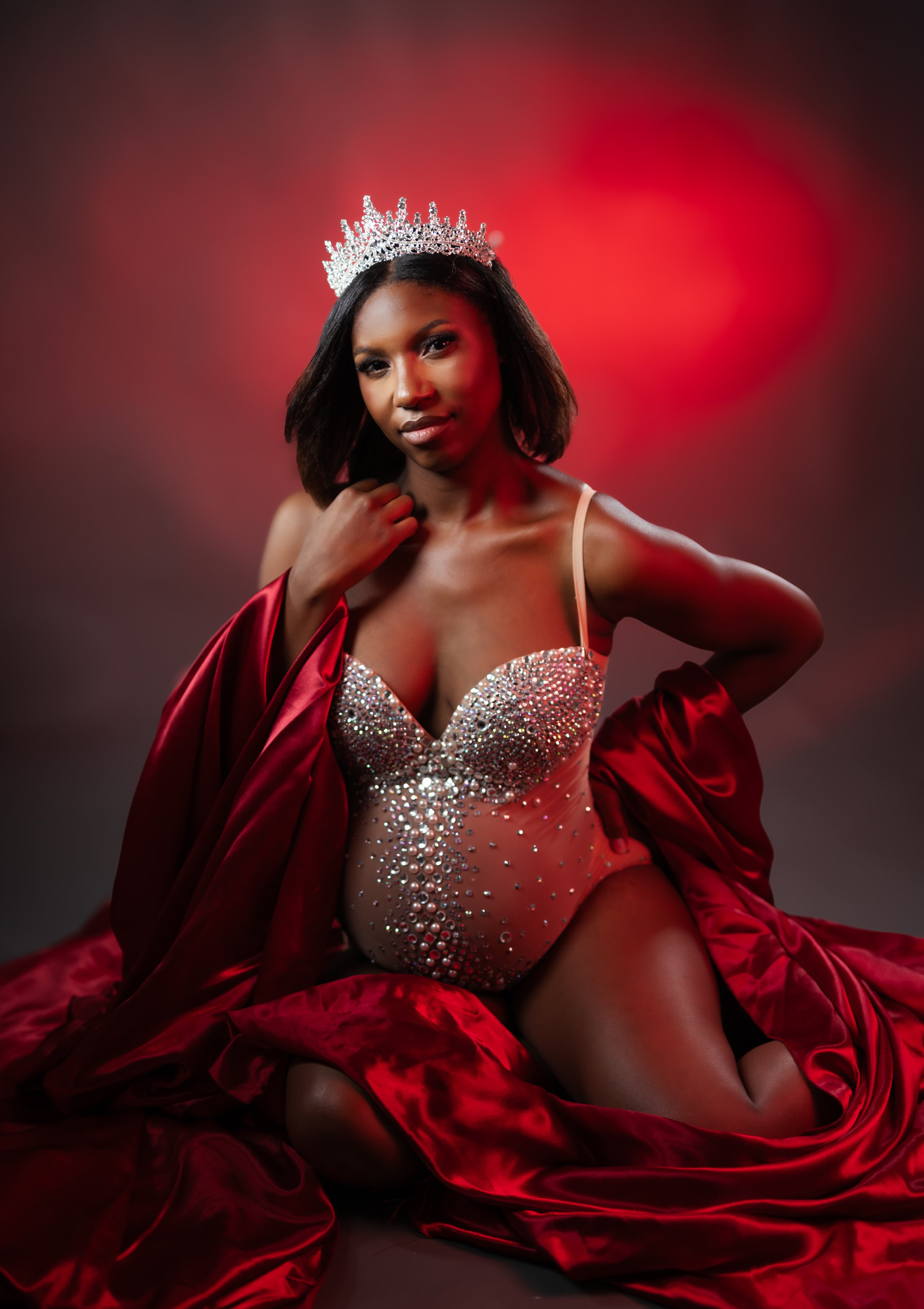 A woman wearing a silver crown and a sparkling bustier with scattered gems, draped in a red satin fabric, seated against a red and black blurred background.