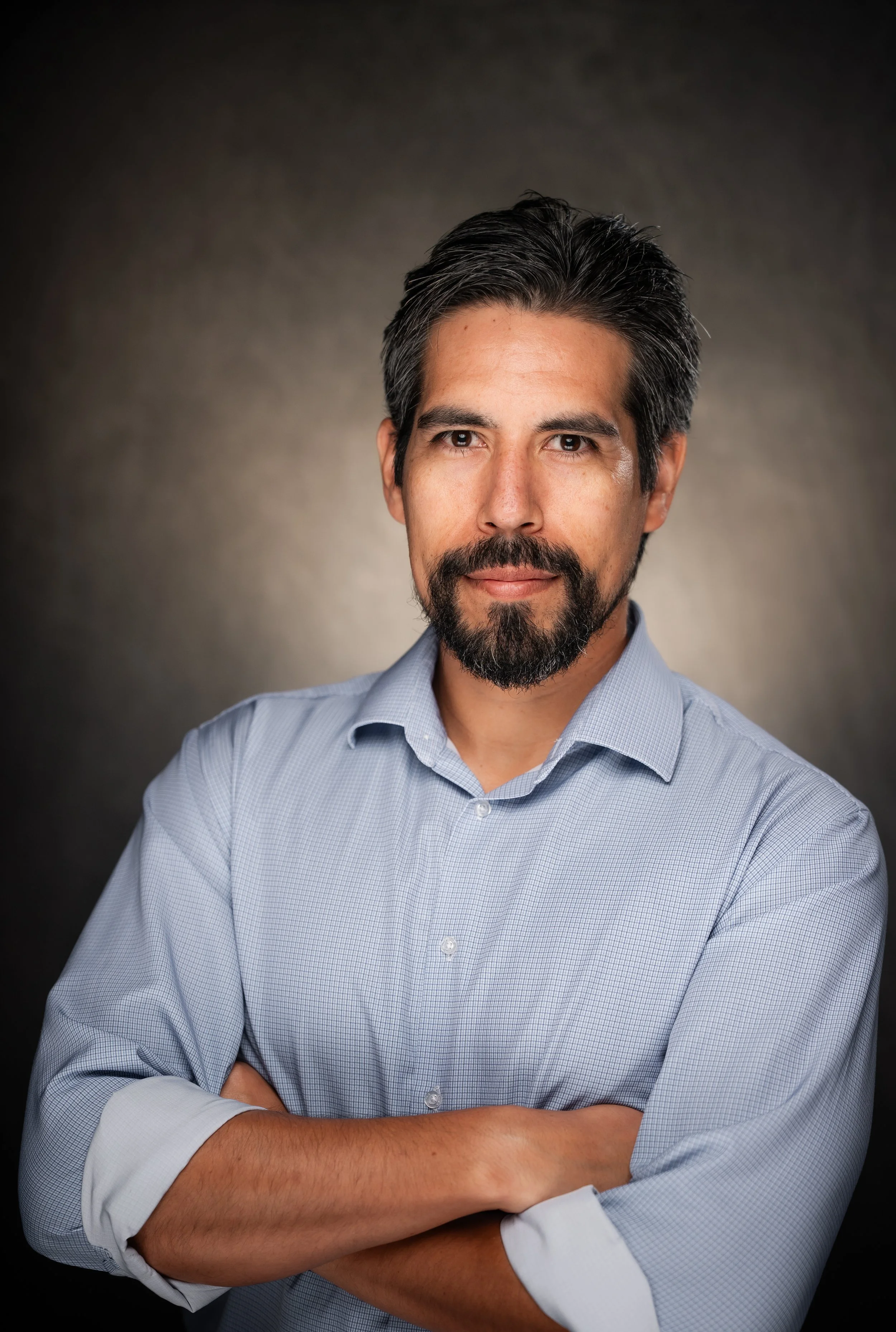 Professional portrait of a bearded man in a light blue shirt with arms crossed.