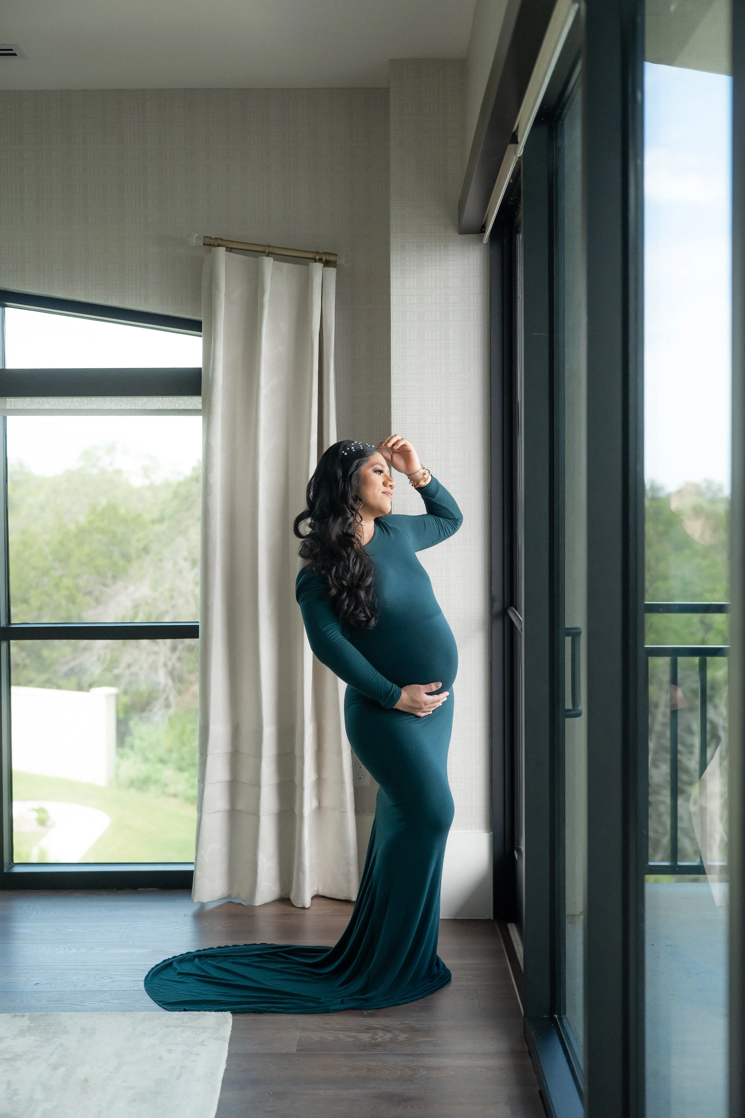 A pregnant woman in a long teal dress standing indoors near large windows with white curtains, looking out and touching her hair with one hand and resting her other hand on her belly, with a green landscape outside.