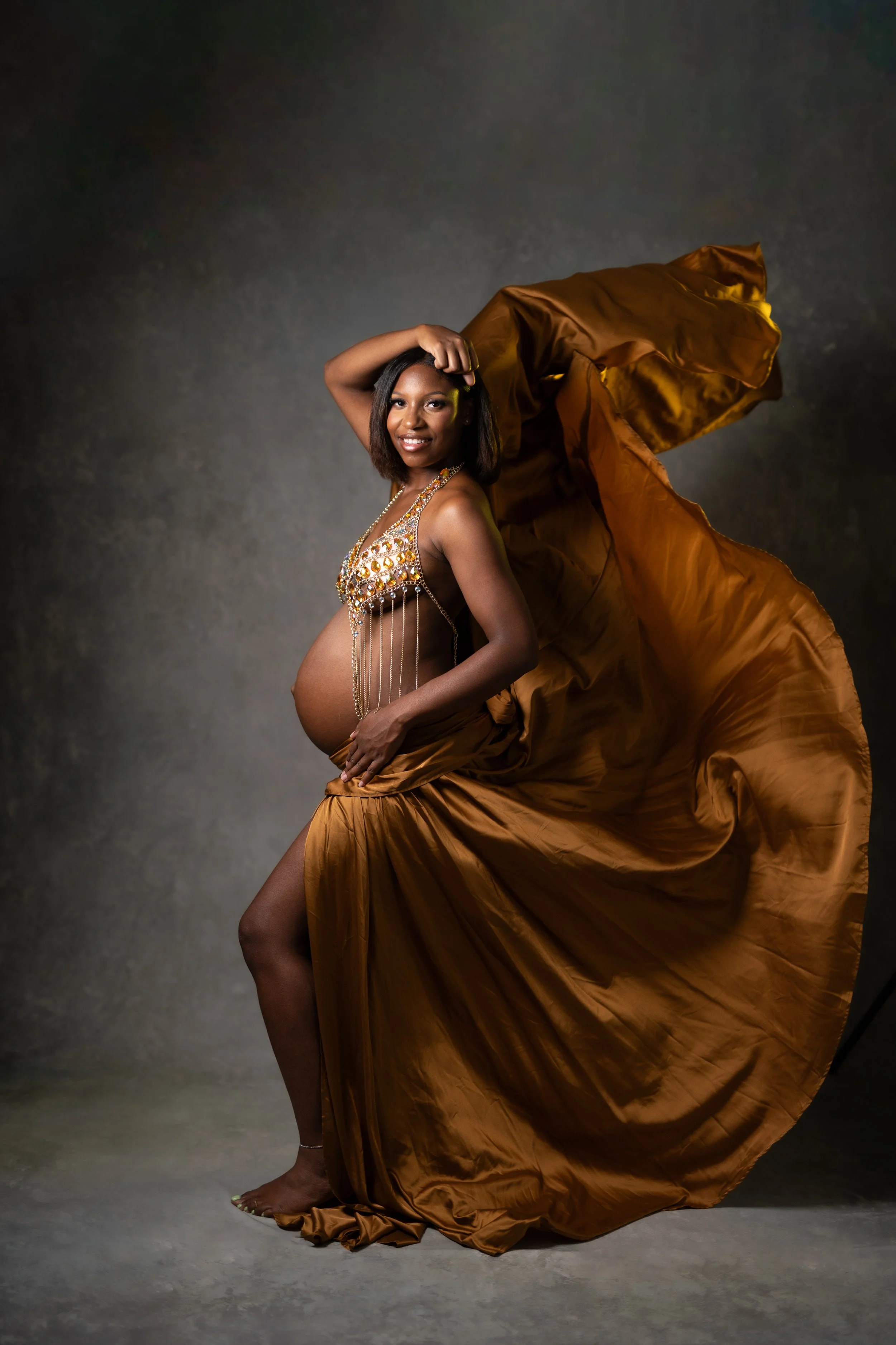 A pregnant woman in a gold dress, smiling, with flowing fabric around her, standing on a textured gray surface with a dark background.