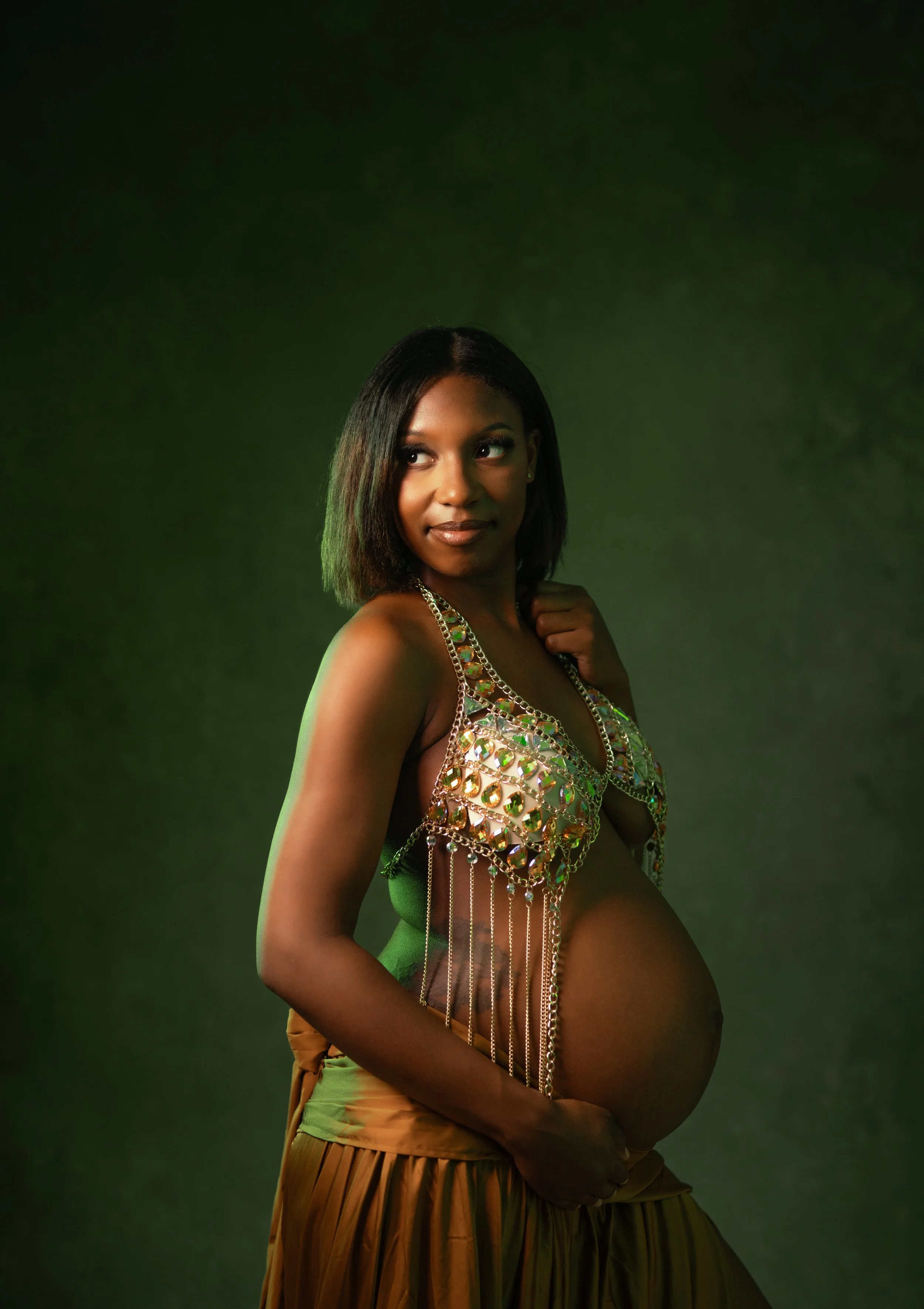 A woman with dark hair and dark skin wearing a decorative top made of iridescent crystals and beads, with loose brown pants, posing against a dark green background.