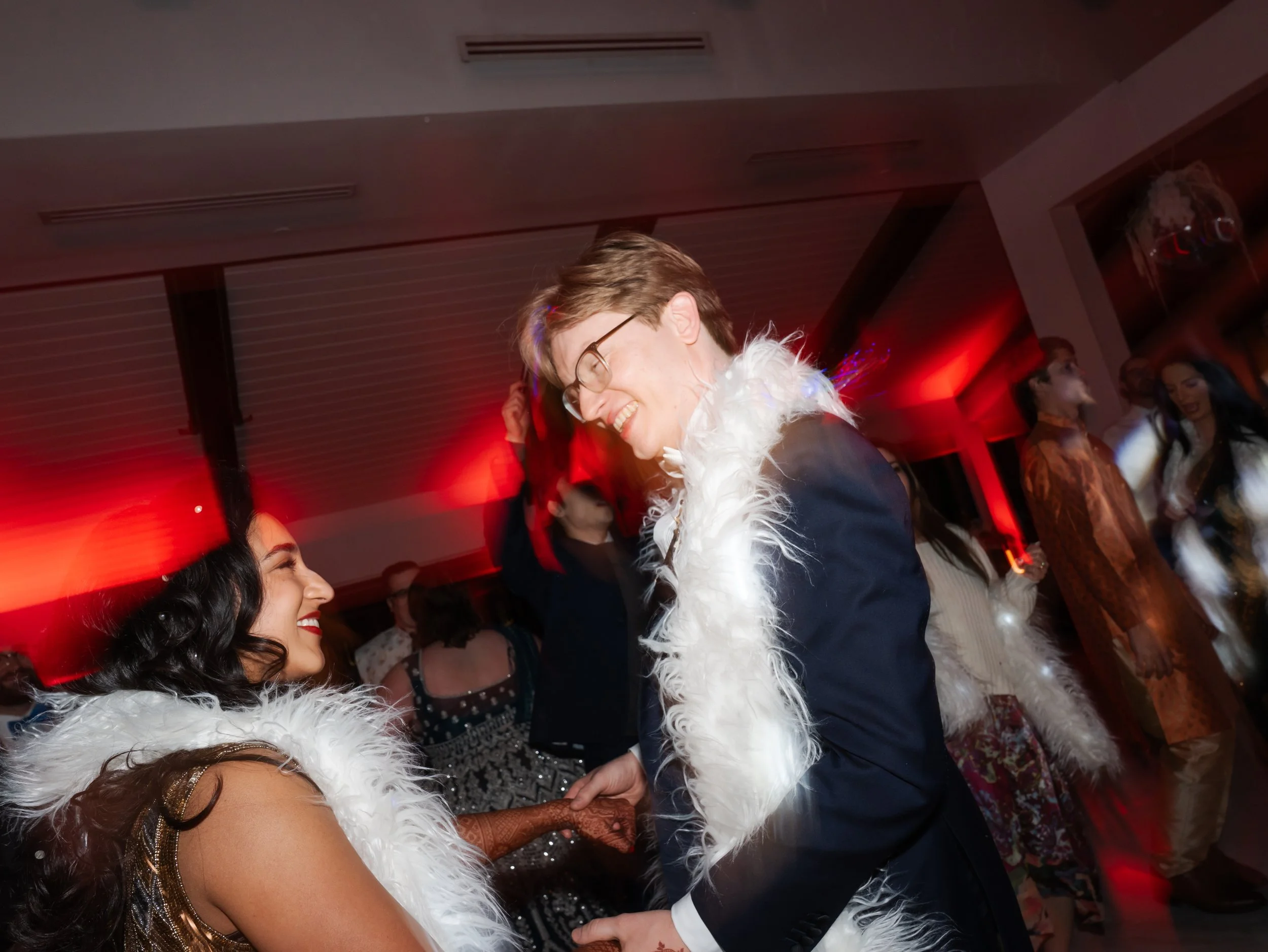 A bride and groom dancing at a party, smiling and holding hands, with everyone dressed in festive attire and red lighting in the background during their New Years Eve wedding at the Videre Estate.