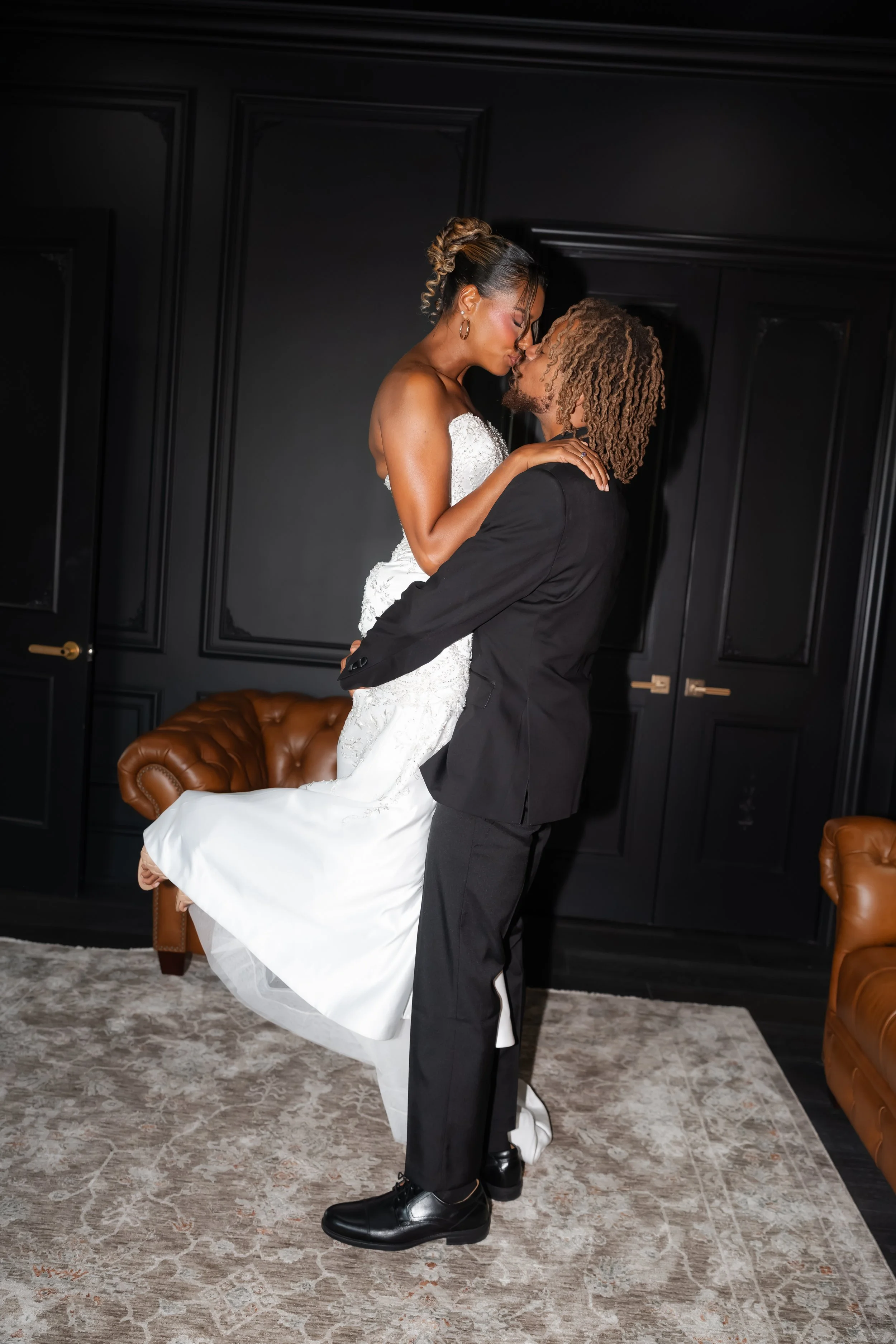 Bride and groom sharing a kiss beside a leather sofa in a dark, paneled room.
