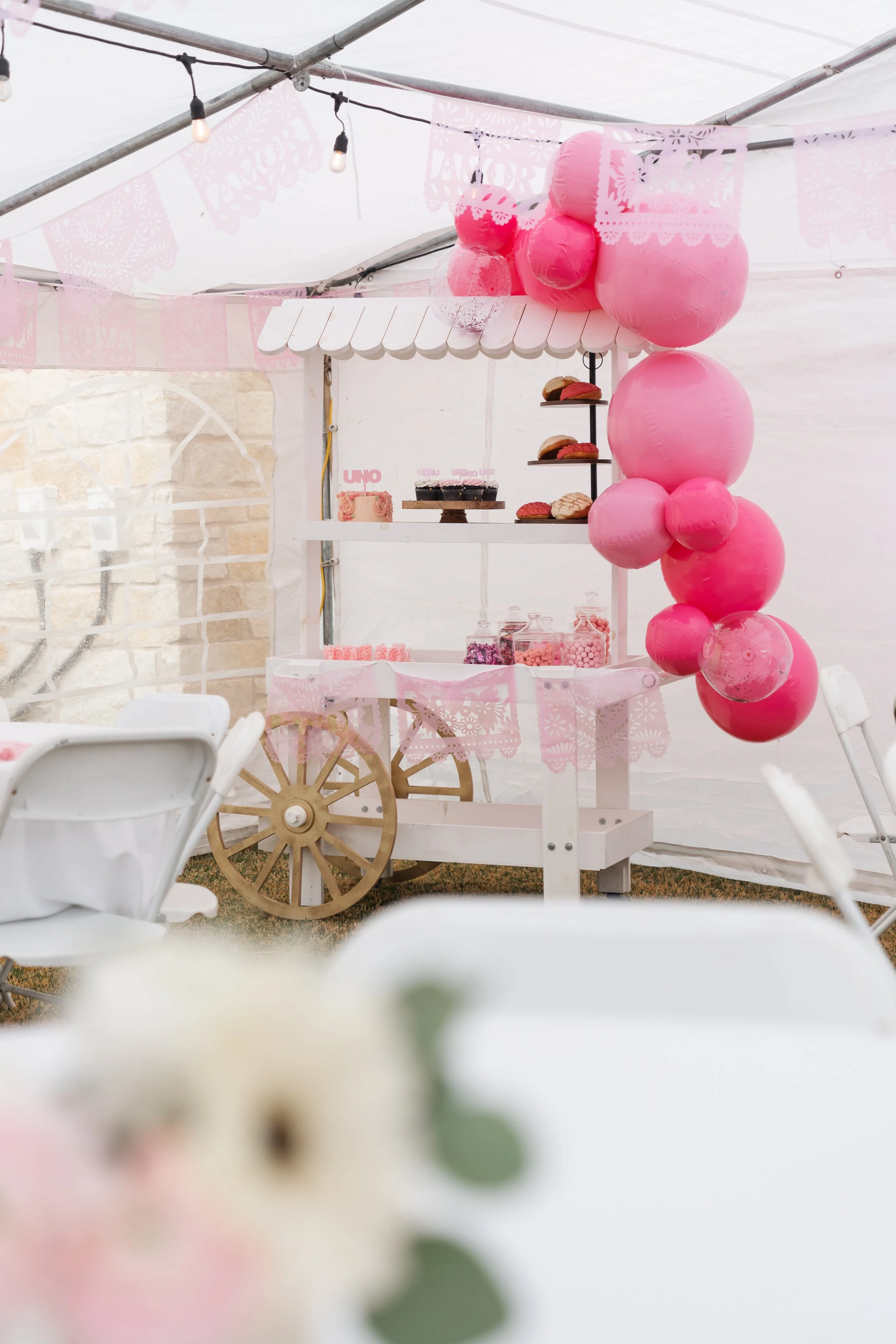 Decorated party booth with pink balloons, a white cart with awning, cupcakes, candies, and a pink themed dessert setup in a tent with string lights.