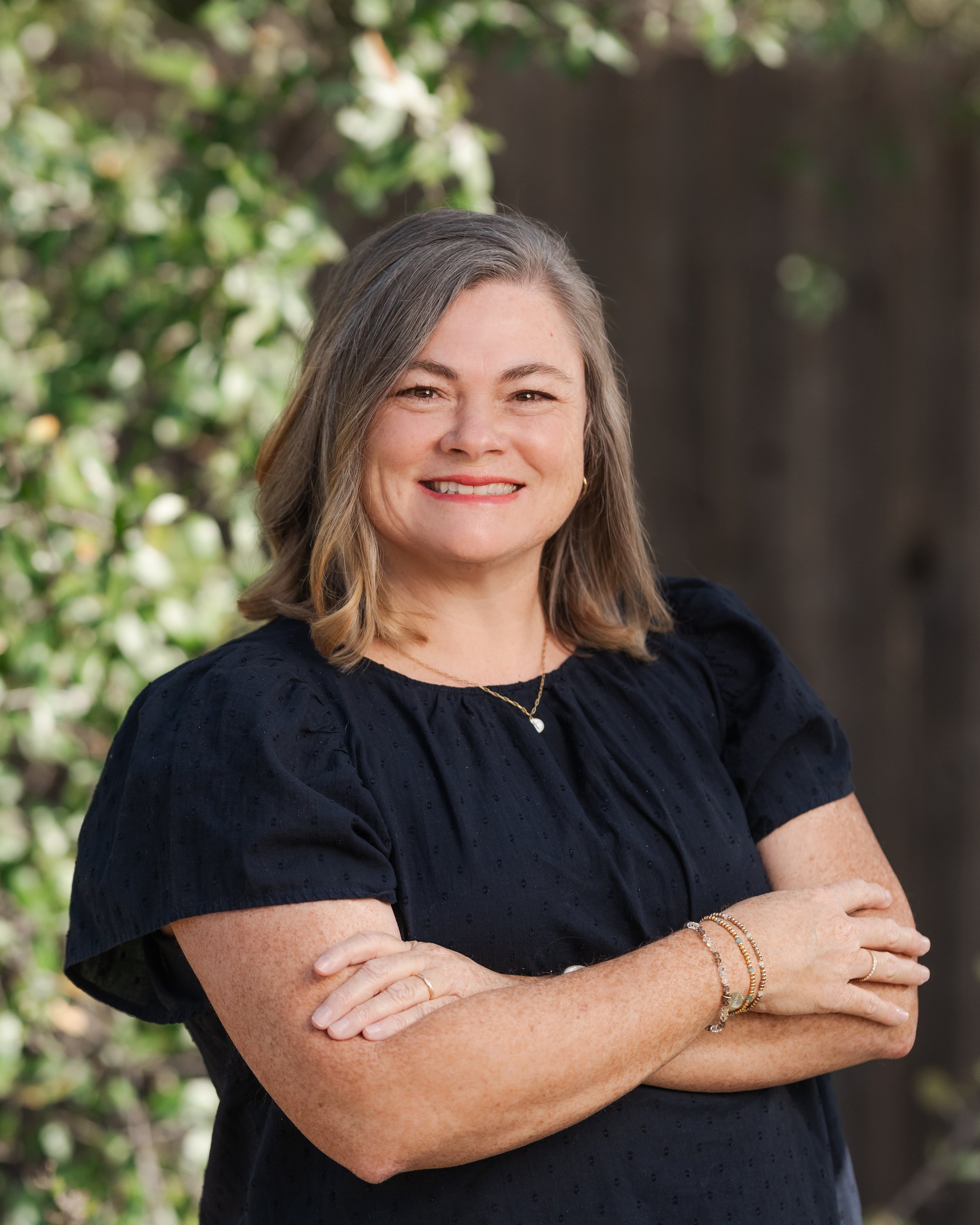 A woman with shoulder-length brown hair, smiling, wearing a black blouse, standing outdoors with green foliage and a wooden fence in the background.