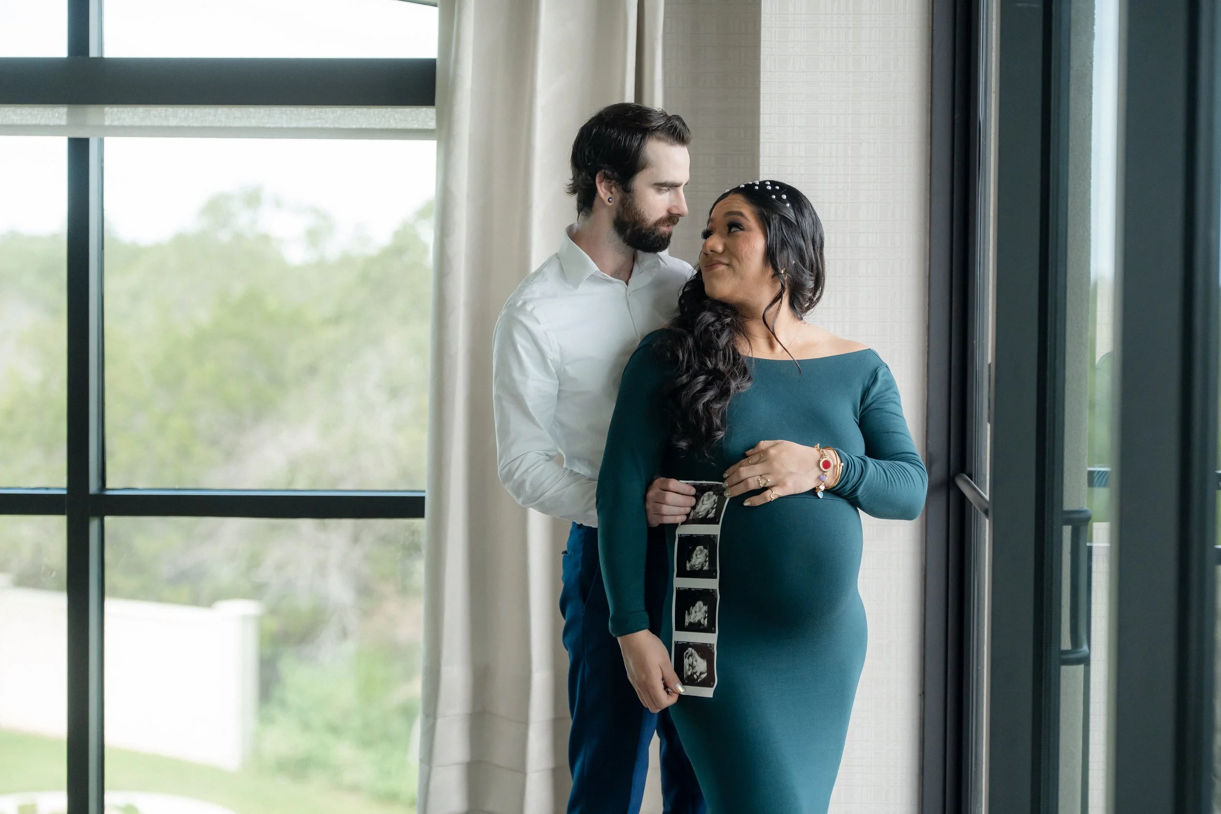 A man and a pregnant woman standing by a large window, looking at each other. The woman is holding ultrasound images. The scene appears to be indoors with a view of trees outside.