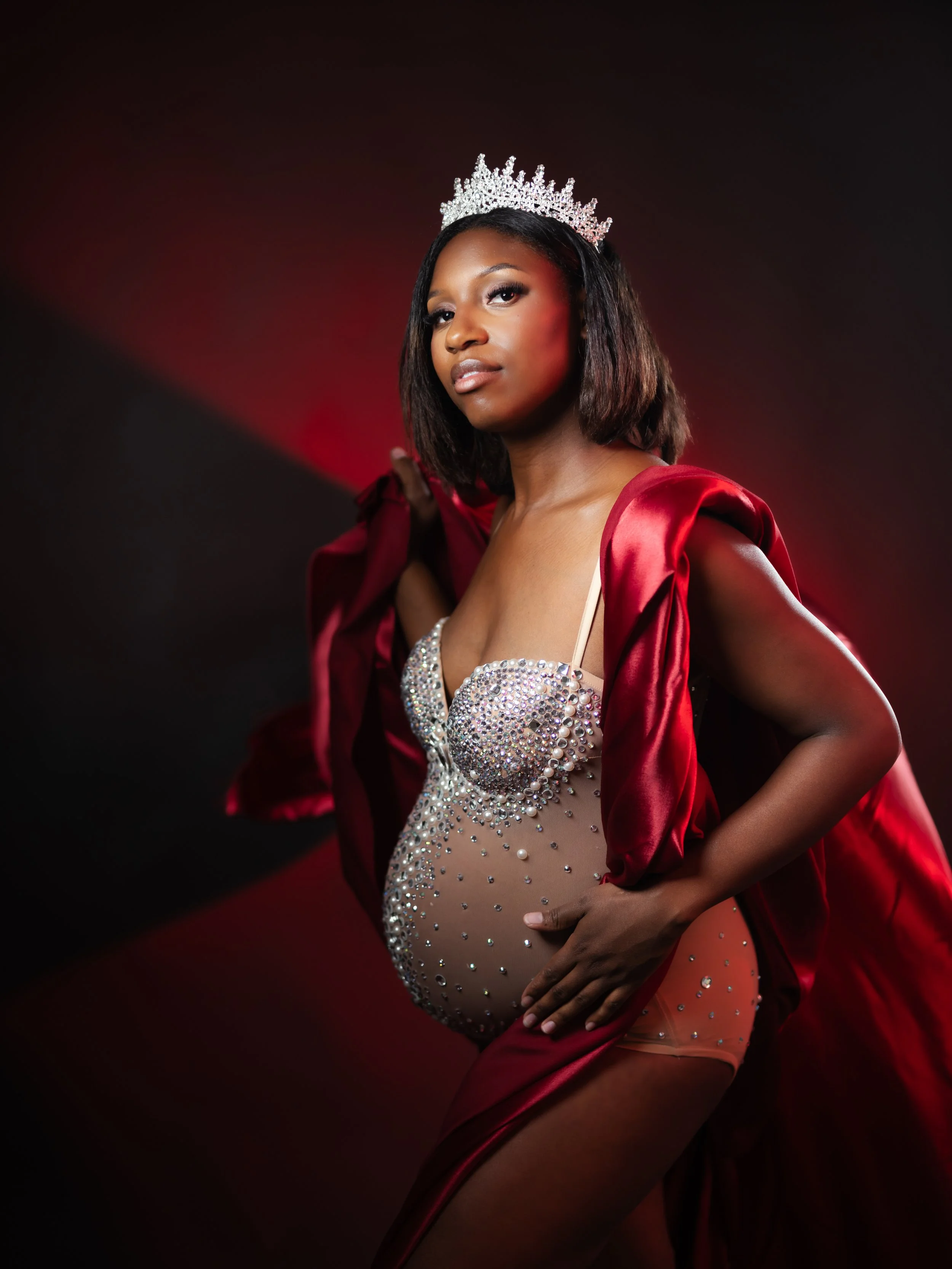 A woman wearing a crown and a red satin robe, posing with her hand on her hip against a dark and red gradient background.