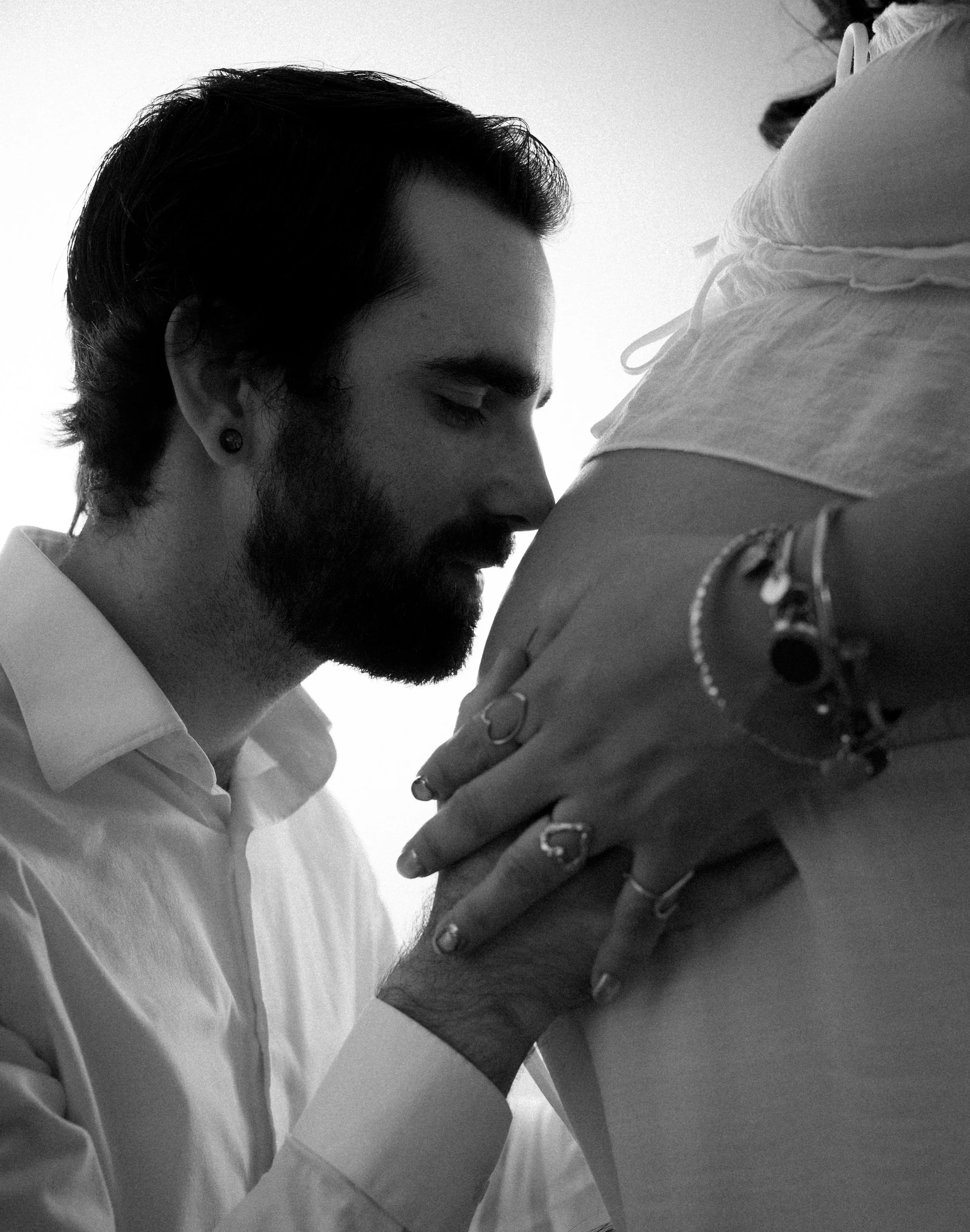 A black and white photo of a man with dark hair and a beard holding a woman close, with their heads bowed together in an intimate moment.