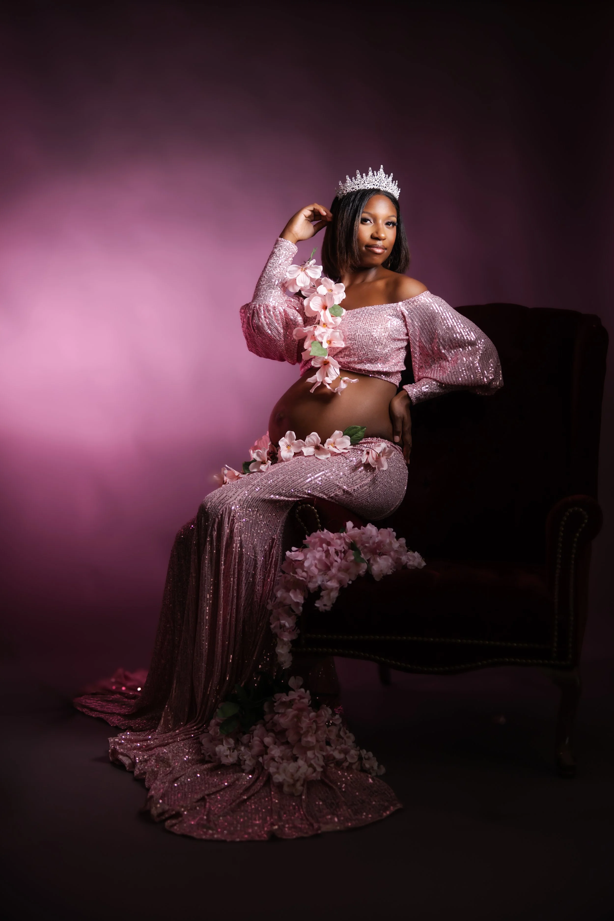 A woman wearing a sparkling pink dress and a silver tiara, sitting on a dark chair surrounded by pink flowers, against a dark gradient background.