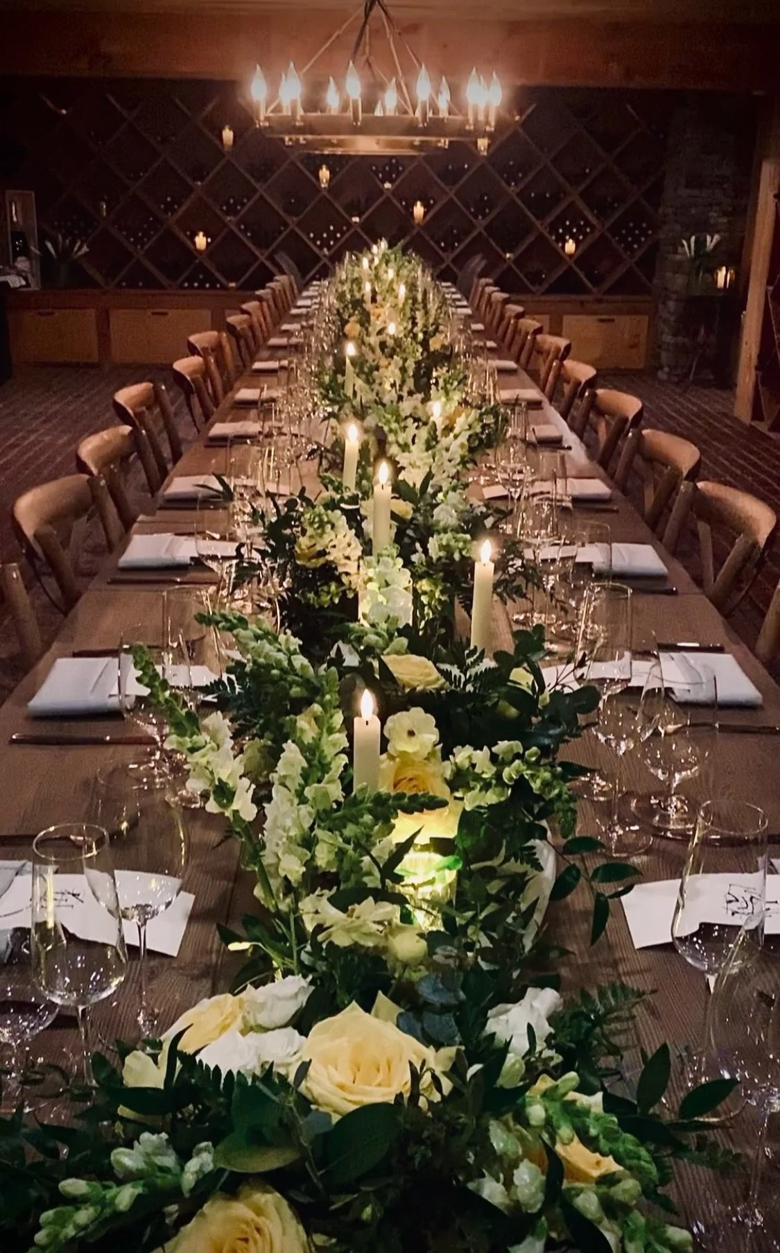 Elegant dining table set for a formal event with candles, white flowers, and glassware, in a rustic wine cellar or vineyard interior.