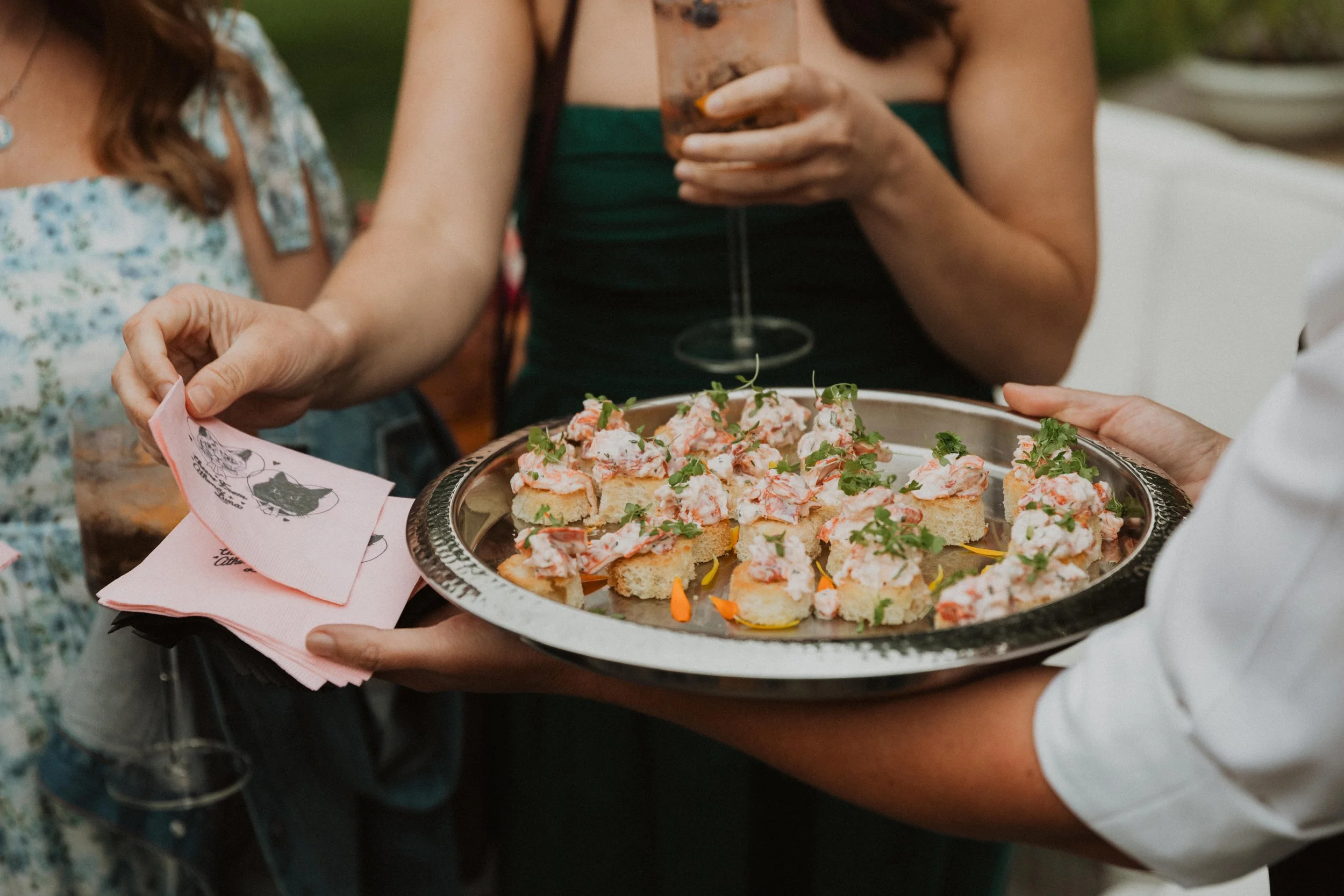 Person serving a platter of shrimp canapés topped with herbs and garnishes at a social gathering.