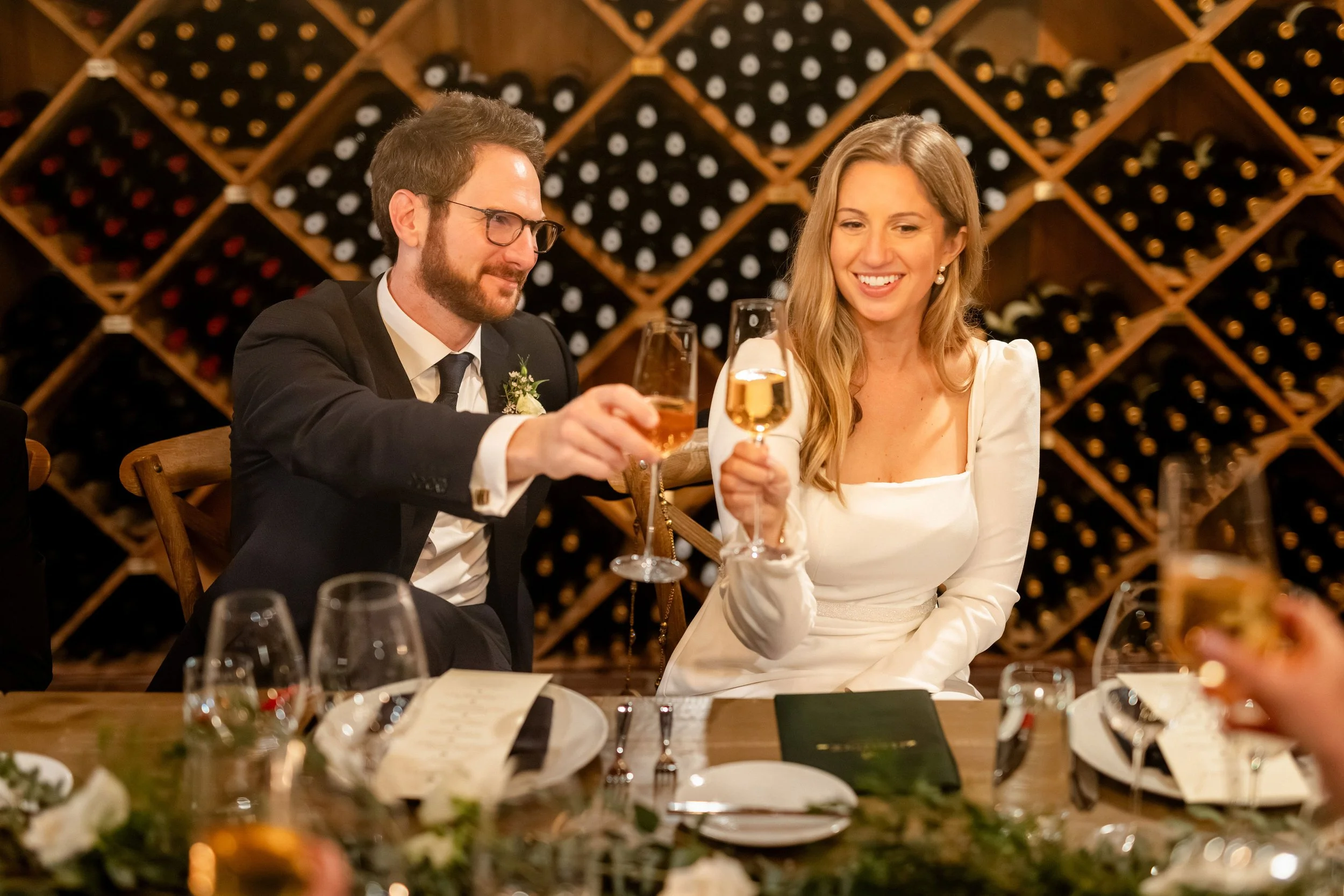 A man and woman at a celebration raising glasses, with wine racks in the background.