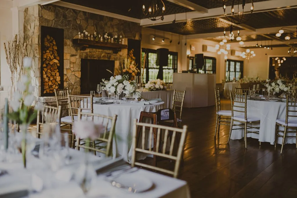 Elegant event space with round tables decorated with white tablecloths and floral centerpieces, gold chairs, a stone fireplace, and warm lighting.