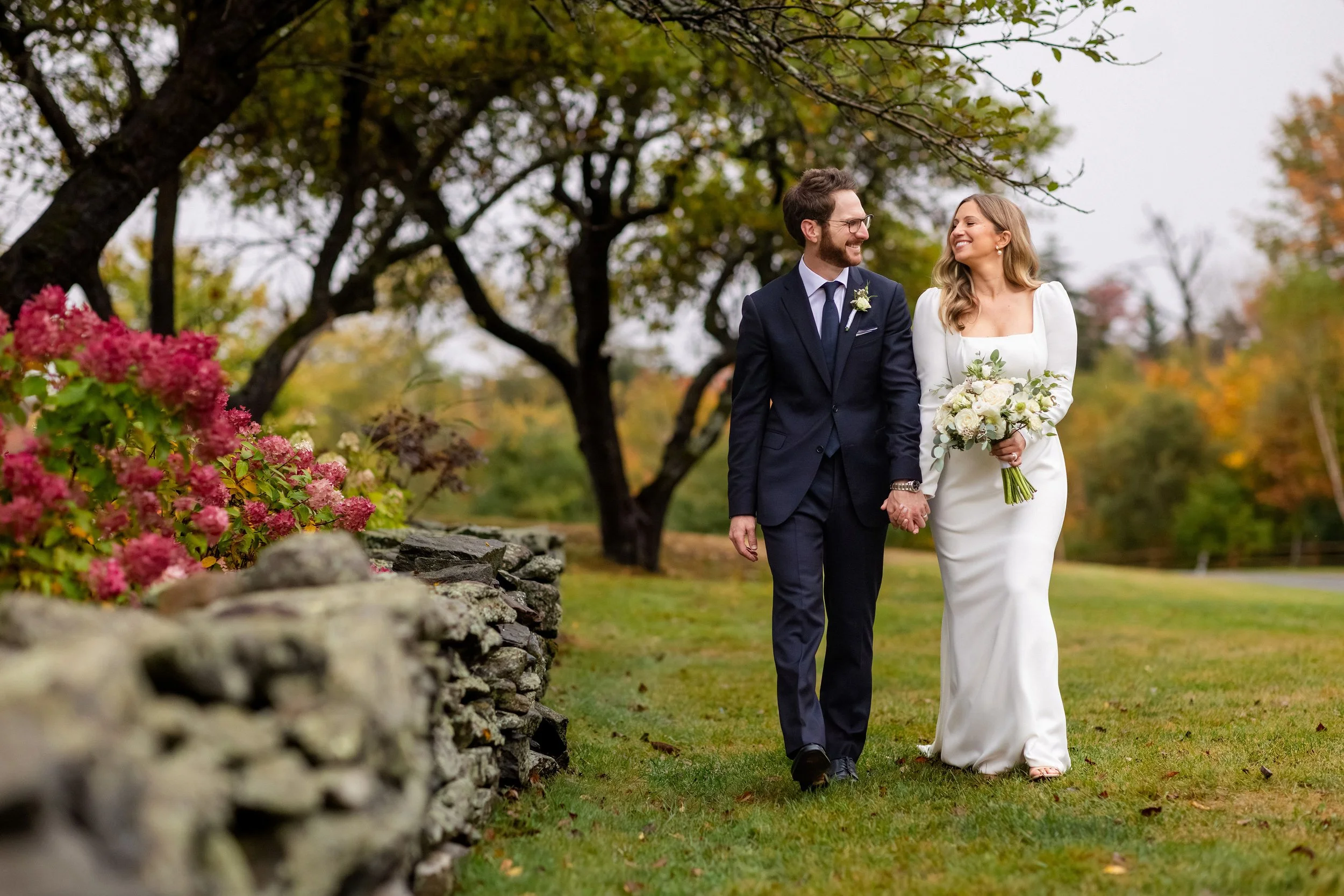 A newlywed couple walking outdoors in a park with colorful autumn trees in the background. The bride is wearing a white wedding dress and holding a bouquet of white flowers, while the groom is dressed in a dark suit with a boutonniere. They are holding hands and smiling at each other.