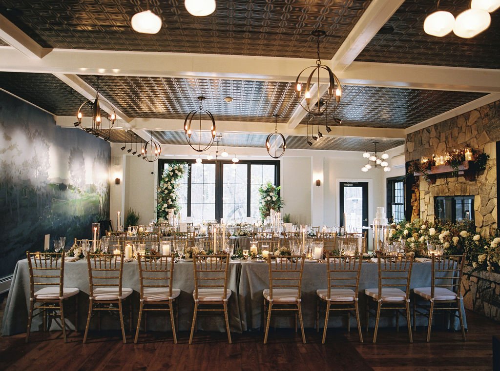 Elegant indoor wedding reception setup with a long table, gold chairs, floral centerpieces, candles, and soft lighting.