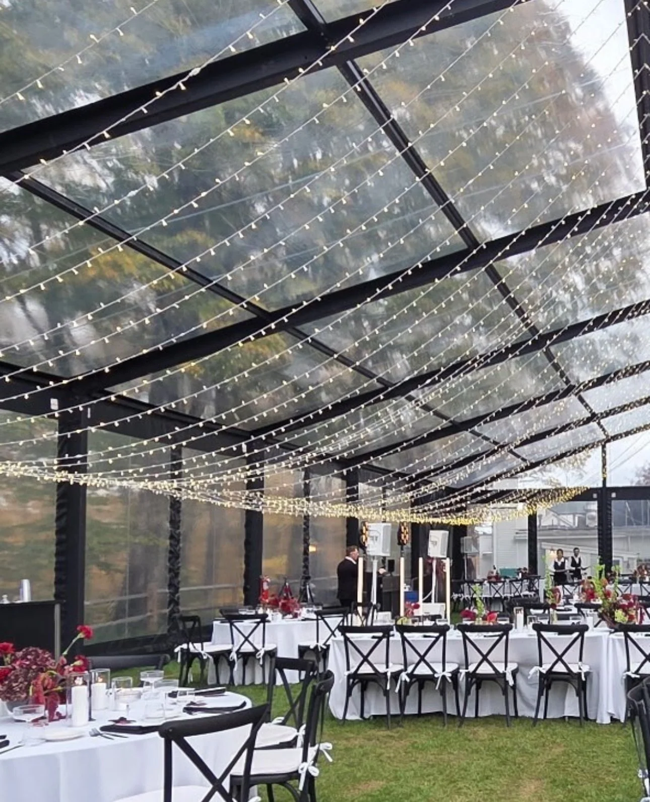 Decorated outdoor dining area with tables, chairs, and string lights under a transparent roof.
