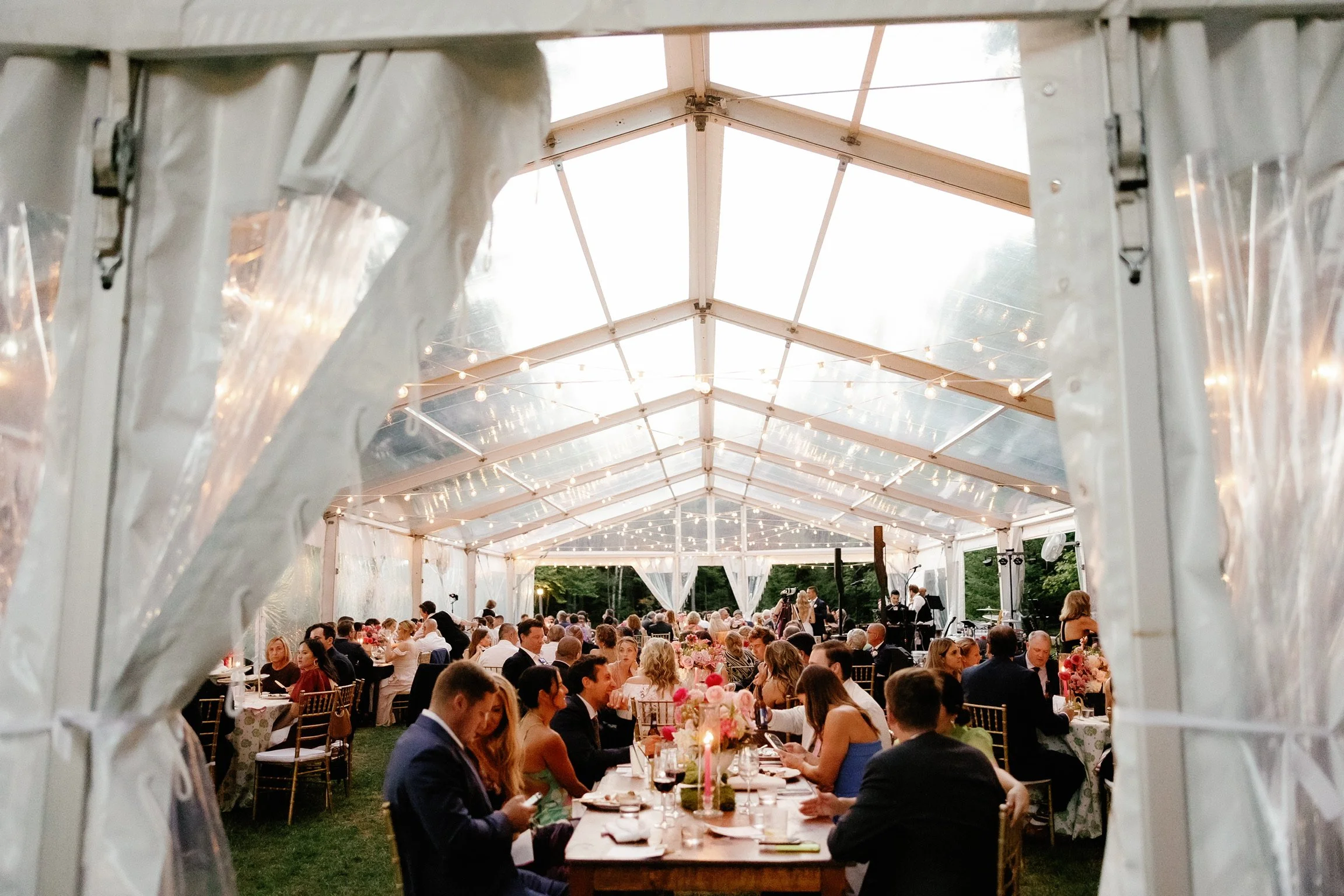 Inside a large white tent with string lights, guests in formal attire are seated at long tables decorated with pink and white flower centerpieces during a wedding reception or celebration.