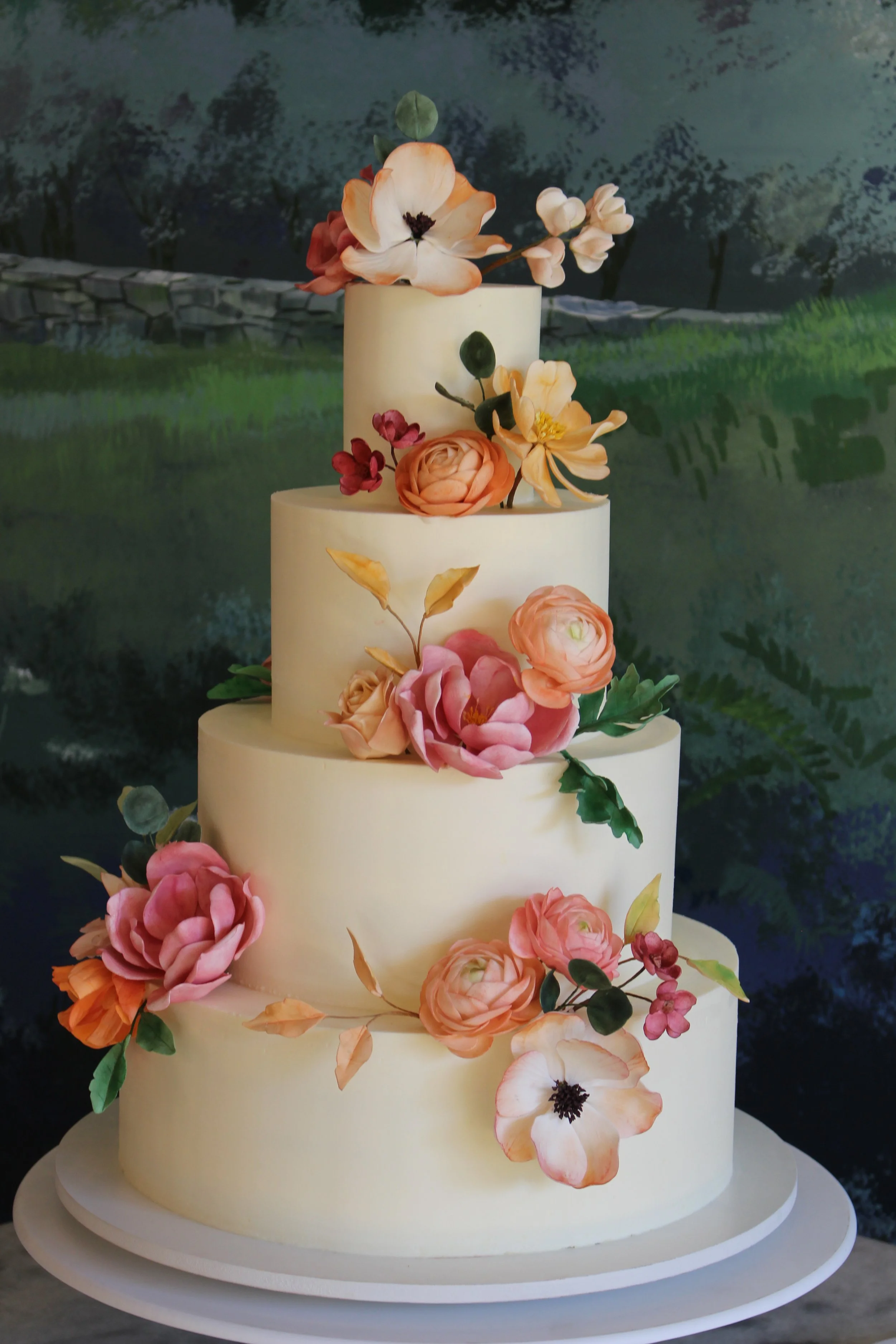 A four-tiered white wedding cake decorated with pink, peach, and cream-colored flowers and greenery, set against a scenic outdoor backdrop.