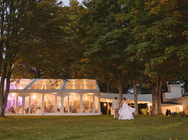 Outdoor wedding reception tent with illuminated interior, surrounded by trees, with a bride and groom walking on grass in front.