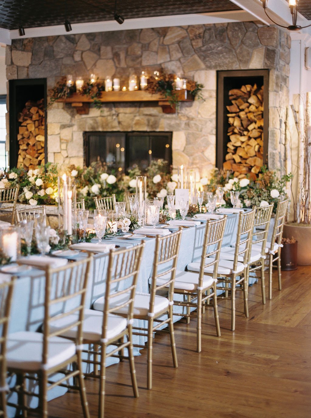 Elegant indoor dining table setup with gold chairs, floral arrangements, candles, and a stone fireplace with wood storage, decorated for an event or wedding.